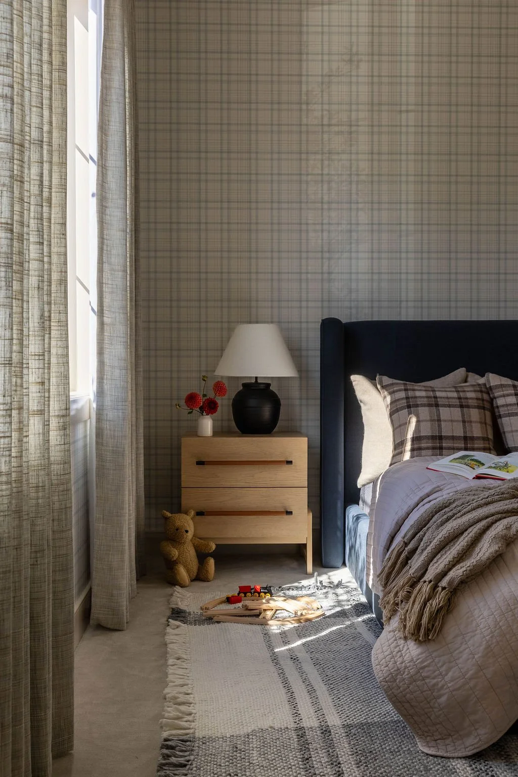 A cozy bedroom corner with a bed, plaid pillows, a beige throw blanket, a wooden nightstand with a white lamp, a small vase with red flowers, a teddy bear, and a train set on a rug near sunlight streaming through a window.