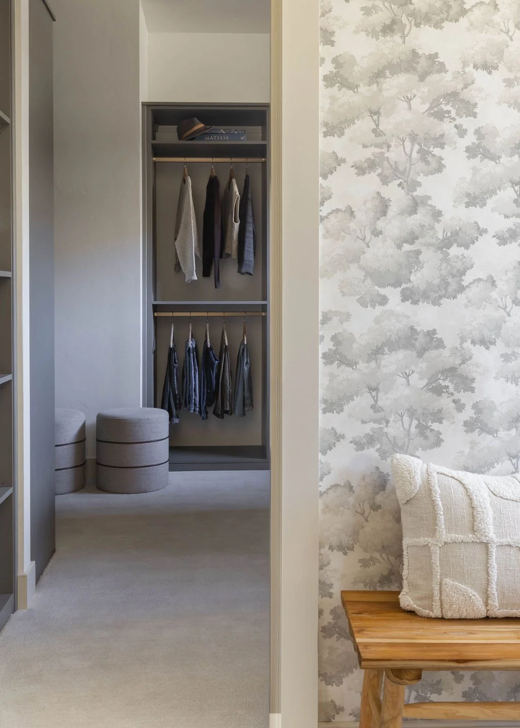 A view into a bedroom closet with hanging clothing and shelves, adjacent to a decorative wall with a bench and cushion.
