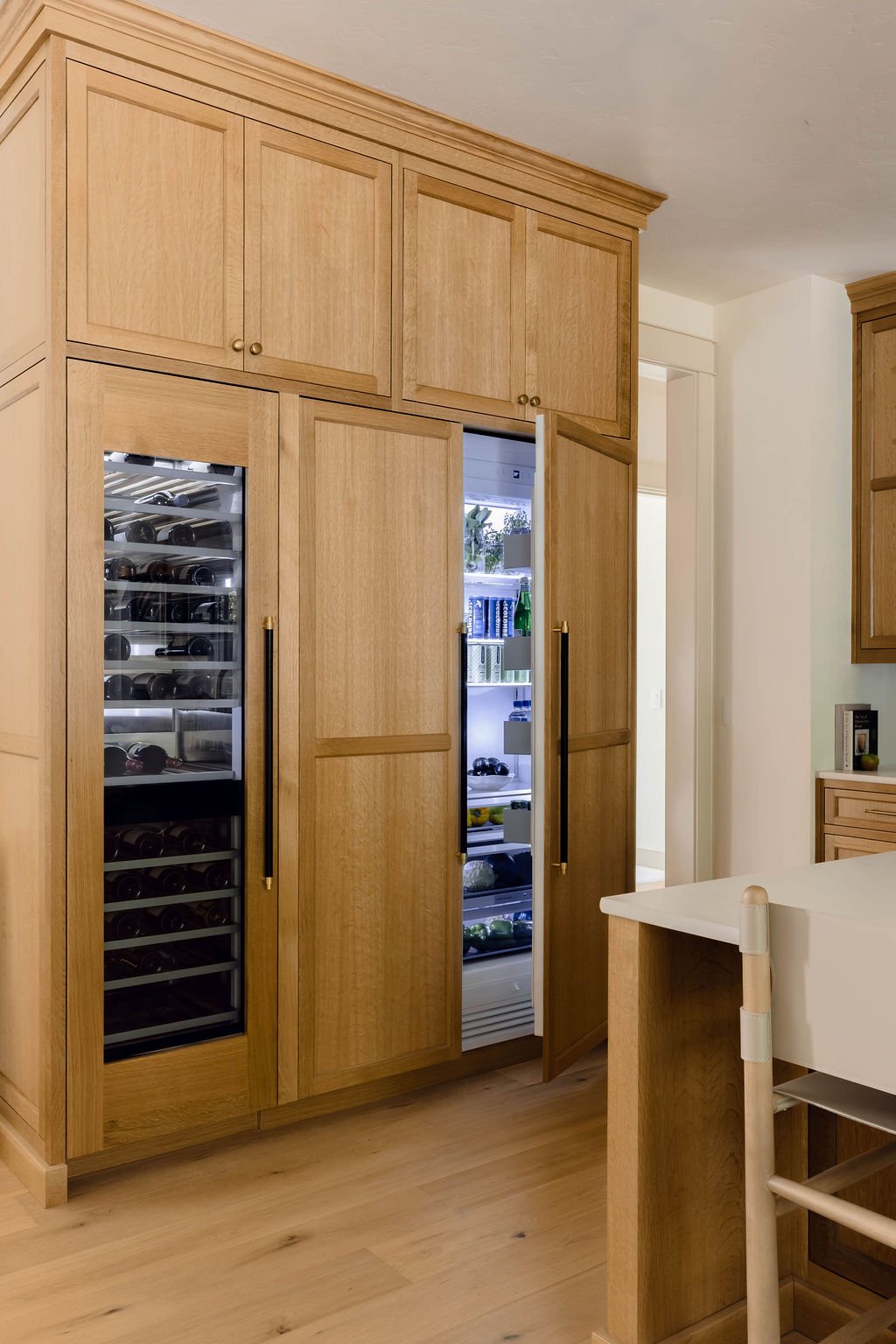 A wooden built-in wine fridge and pantry in a kitchen with cabinets above and an open door showing shelves with bottles and food items.