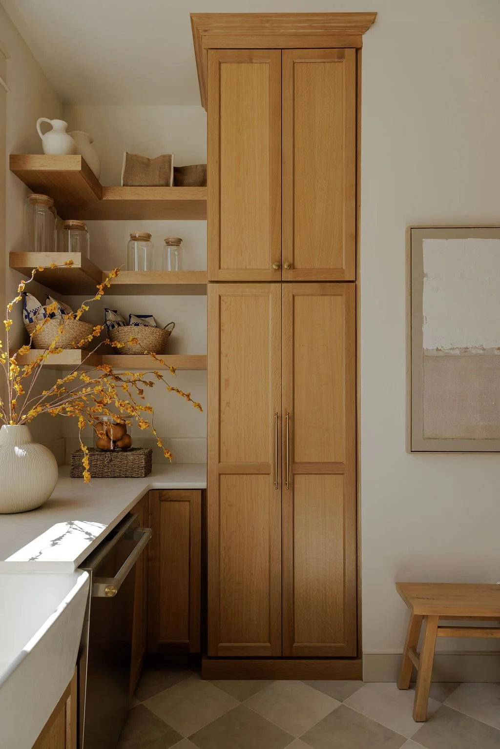Corner of a kitchen with wooden cabinets, open shelves with glass jars and decorative baskets, a white vase with yellow flowers, and a neutral-toned abstract wall art.