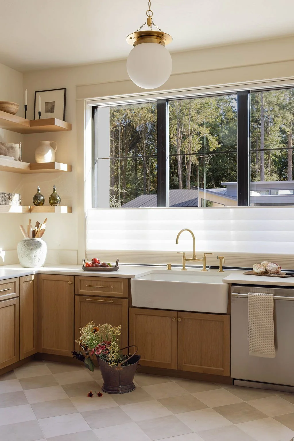 A bright kitchen with large window, wooden cabinets, white farmhouse sink, a bowl of tomatoes, flowers in a bucket, and open shelving with decorative items.