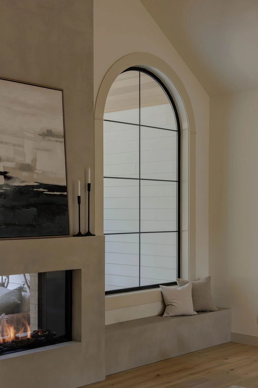 Interior of a modern living room featuring a large arched window with grid pattern, a fireplace with flames, a built-in window bench with pillows, a piece of wall art, and two black candlesticks on a mantle.