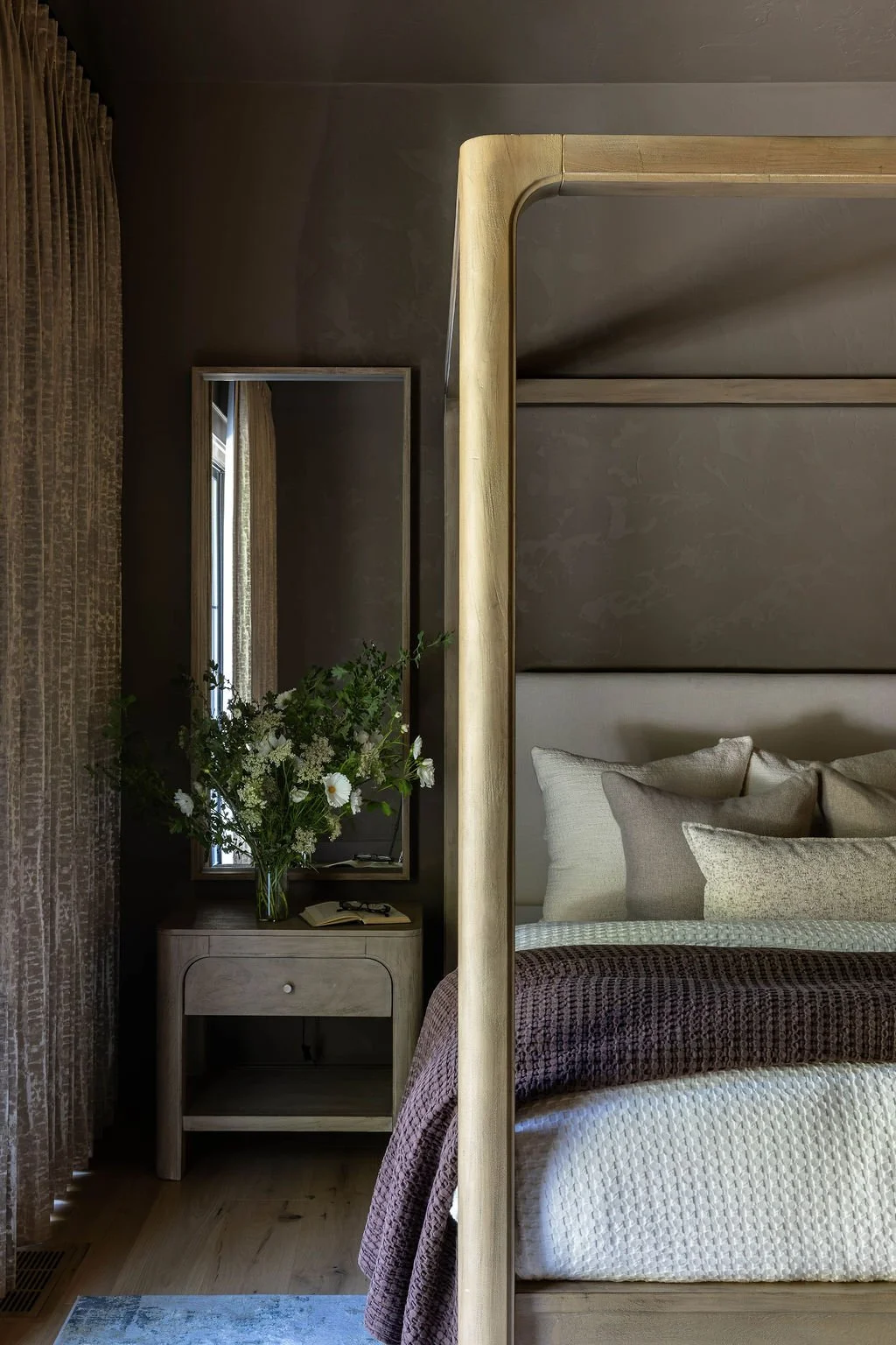 A bedroom corner with a beige upholstered bed, plush pillows, and a wooden canopy. A nightstand with a drawer is next to the bed, holding a large bouquet of white flowers, glasses, and an open book. A mirror reflects a window with beige curtains.