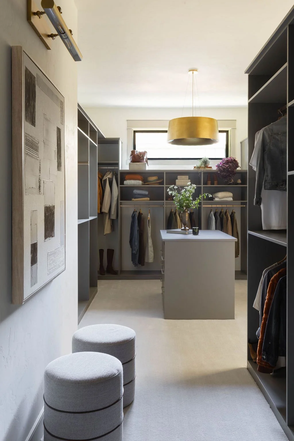 A walk-in closet with gray shelving and hanging clothes, a white island with a vase of flowers, a window with natural light, and two white ottomans in the foreground.