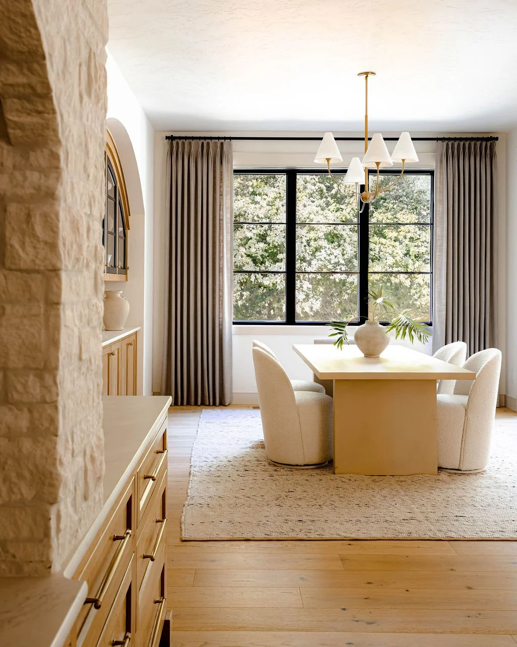 Bright dining room with a beige rectangular table surrounded by plush, cream-colored chairs, a large window with black framing, beige curtains, a modern chandelier, a wooden sideboard, and a patterned area rug, with a vase containing green leafy bran