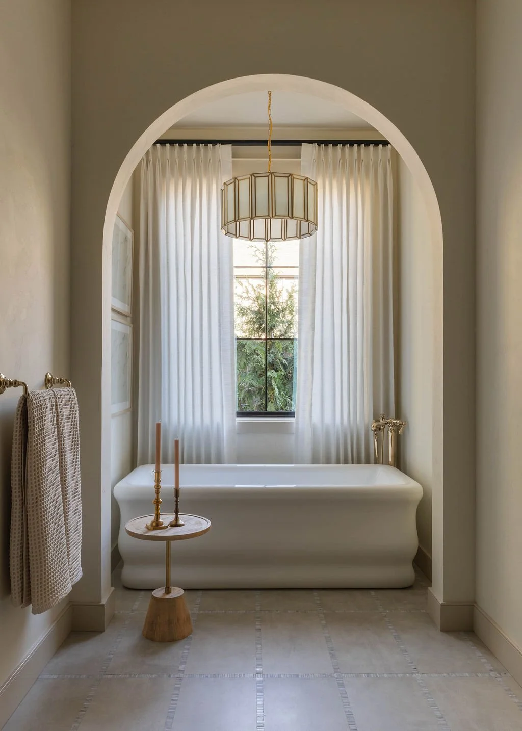 A bathroom with a white bathtub, a window with sheer white curtains, a gold chandelier, and a small wooden table with two candles.