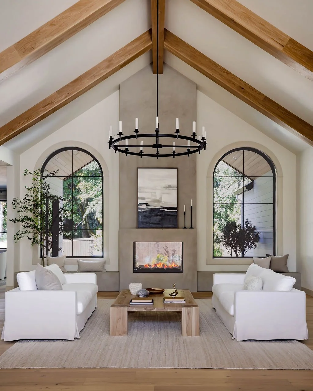 Living room with vaulted ceiling, wooden beams, fireplace, two arched windows, white sofas, wooden coffee table, decorative items, and chandelier.