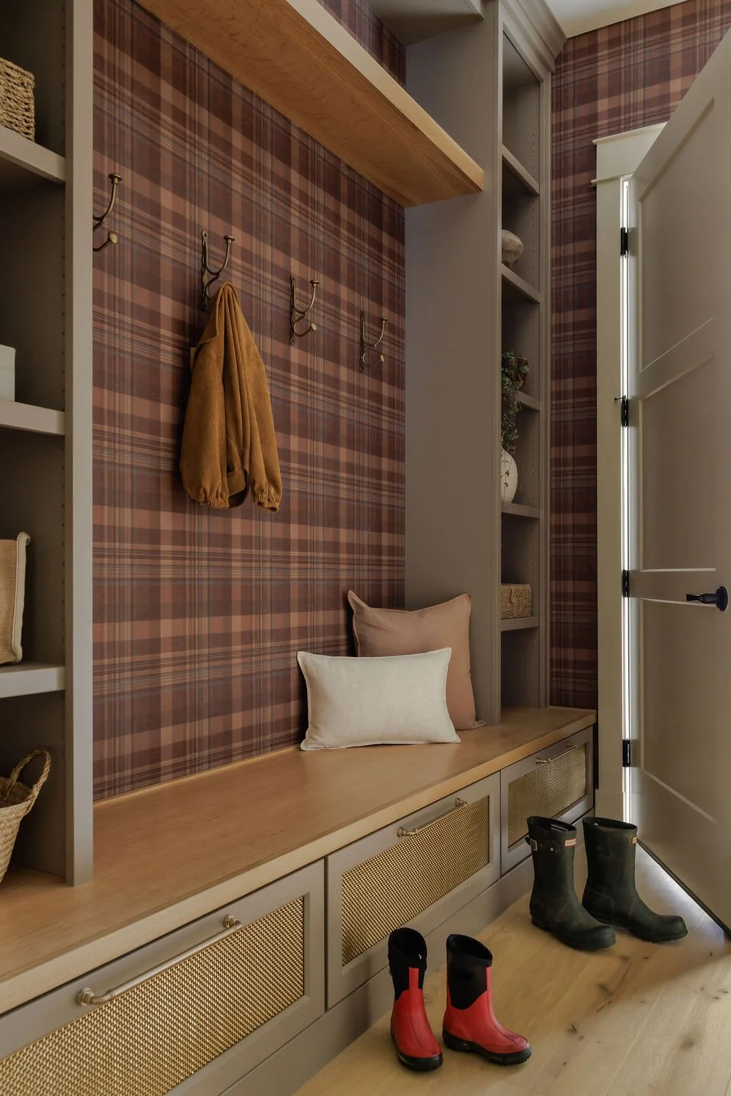 Entryway with a bench, pillows, coat hooks with a coat, shelves with wicker baskets, and rain boots on the floor.