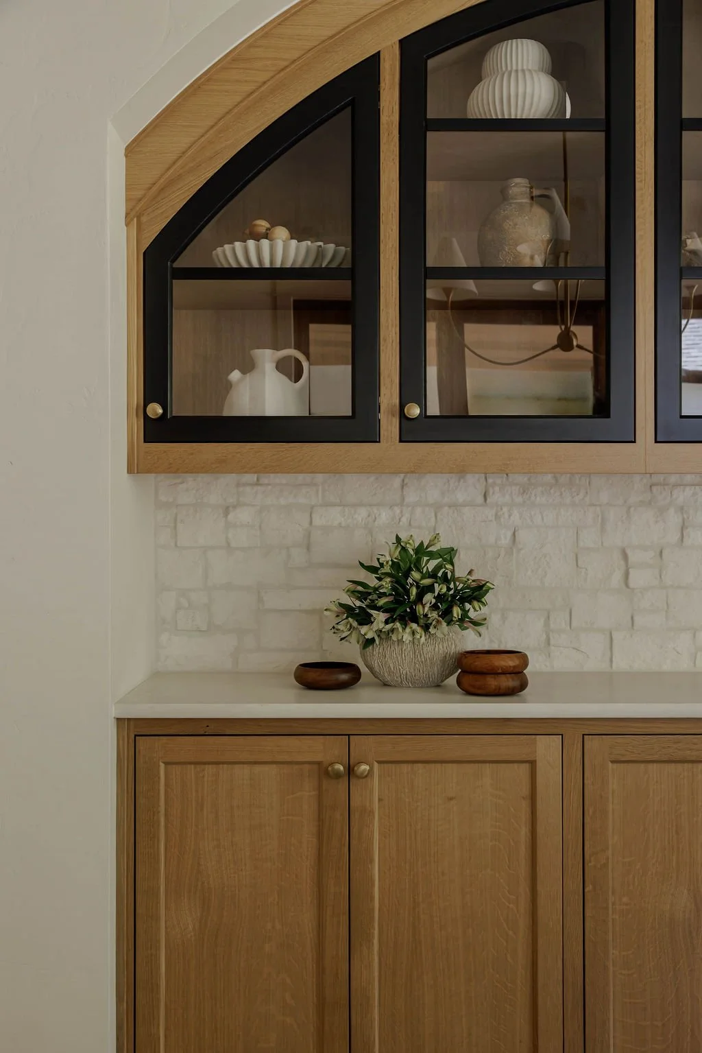 A kitchen cabinet with glass doors displaying decorative vases and bowls, and a countertop with a flower arrangement in a ceramic vase and two wooden bowls.
