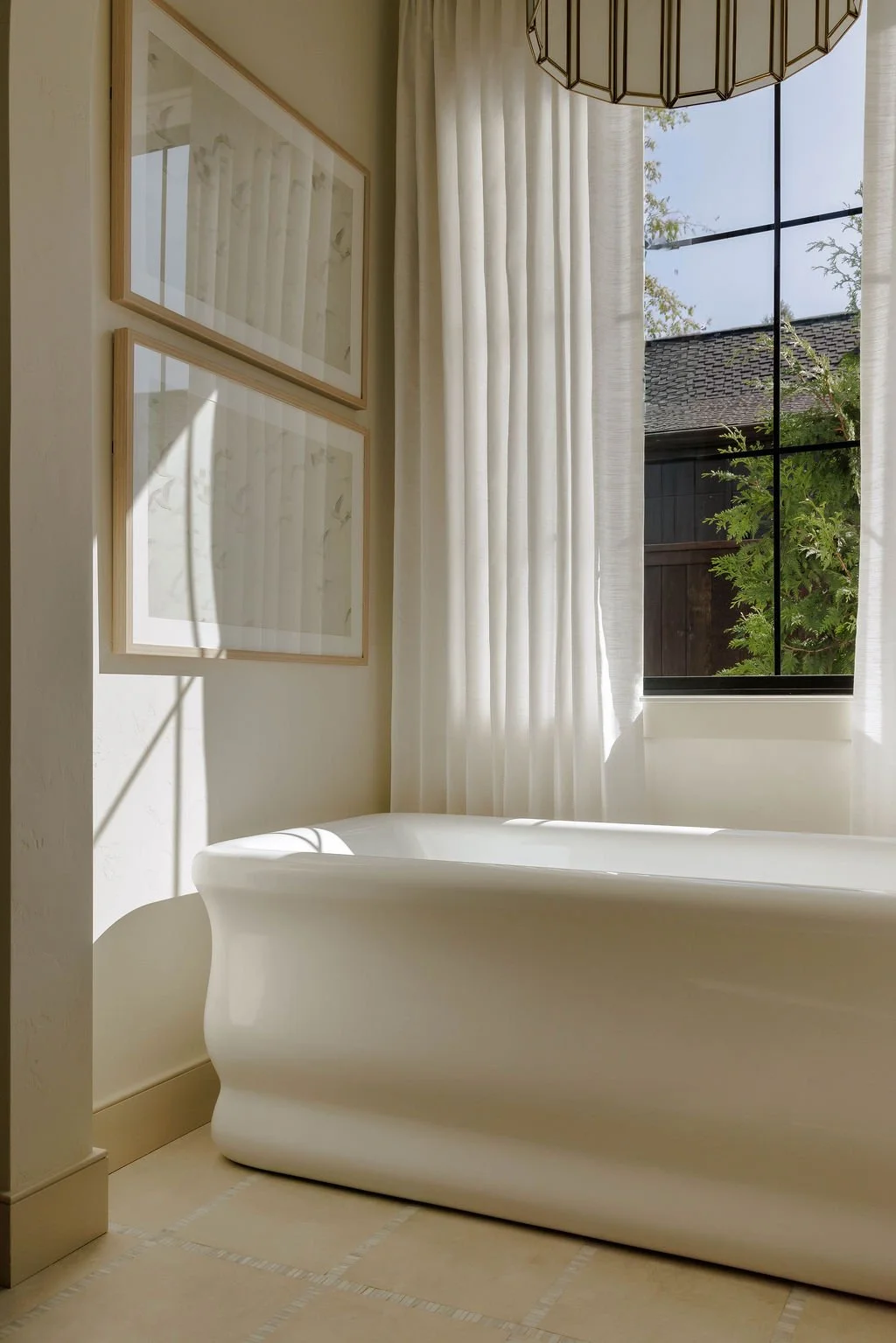 Bright bathroom with a freestanding white bathtub, large window with white curtains, and framed artwork on the wall.
