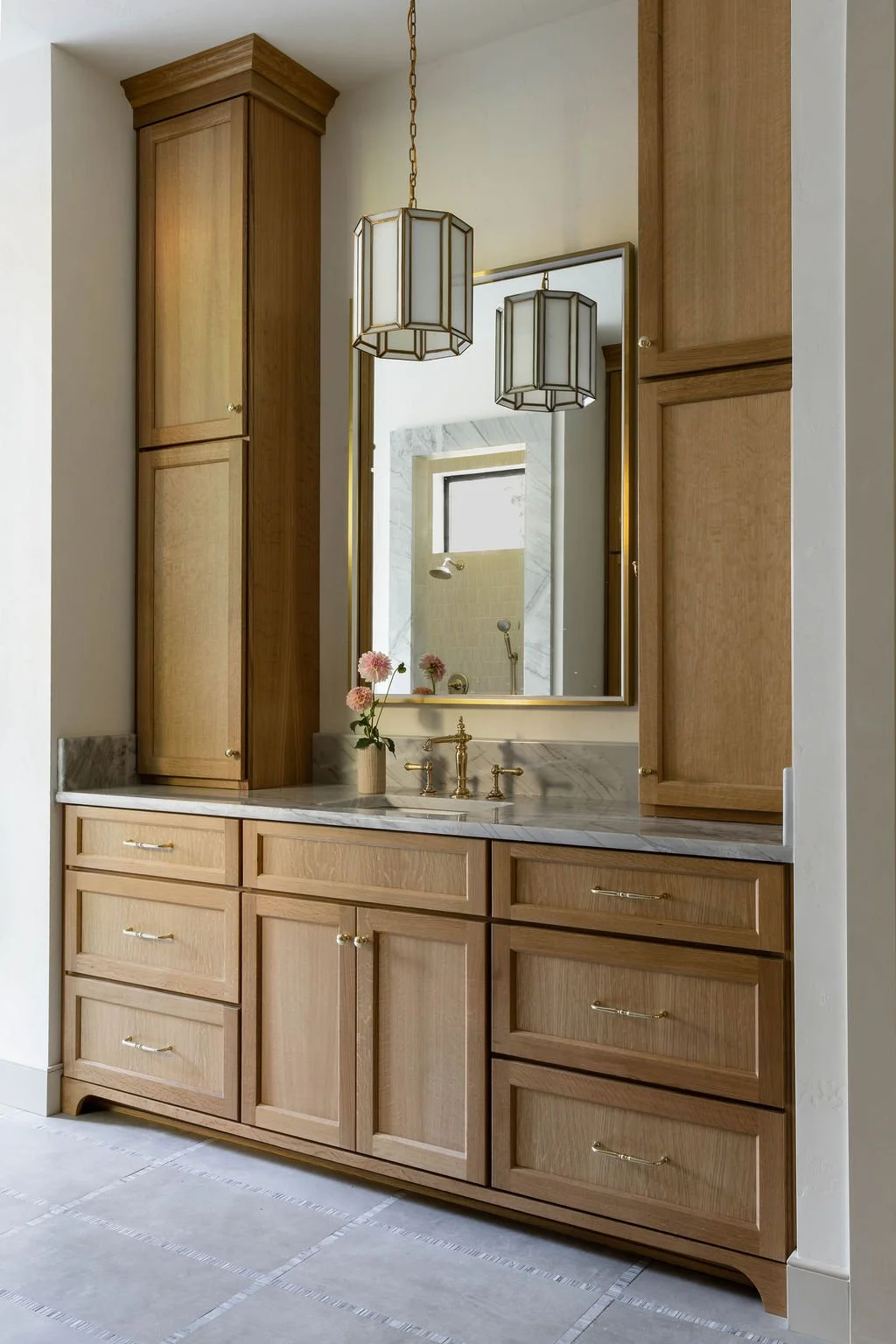 Wood bathroom vanity with marble countertop, large mirror, hanging pendant lights, and a small vase with pink flowers.