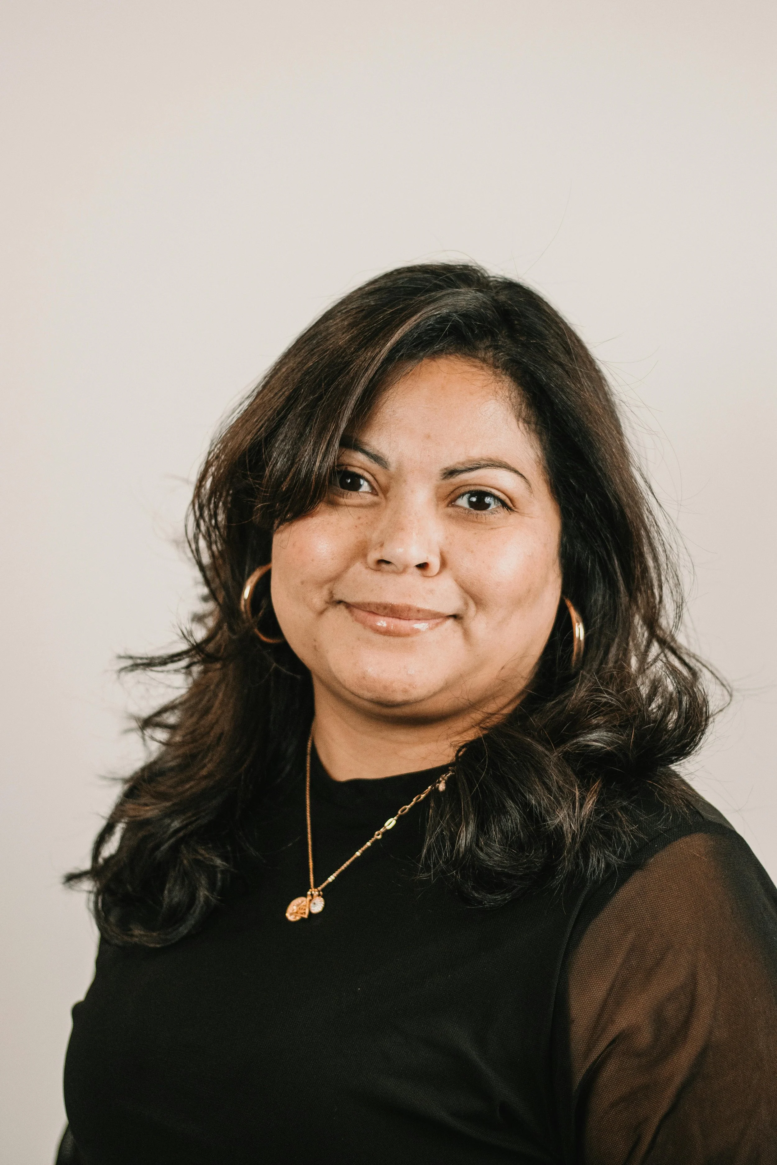 a woman with long dark hair wears a black top and gold jewelry, smiling softly at the camera against a neutral background