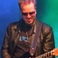 a man wearing sunglasses and a black leather jacket plays an electric guitar on stage under green lighting
