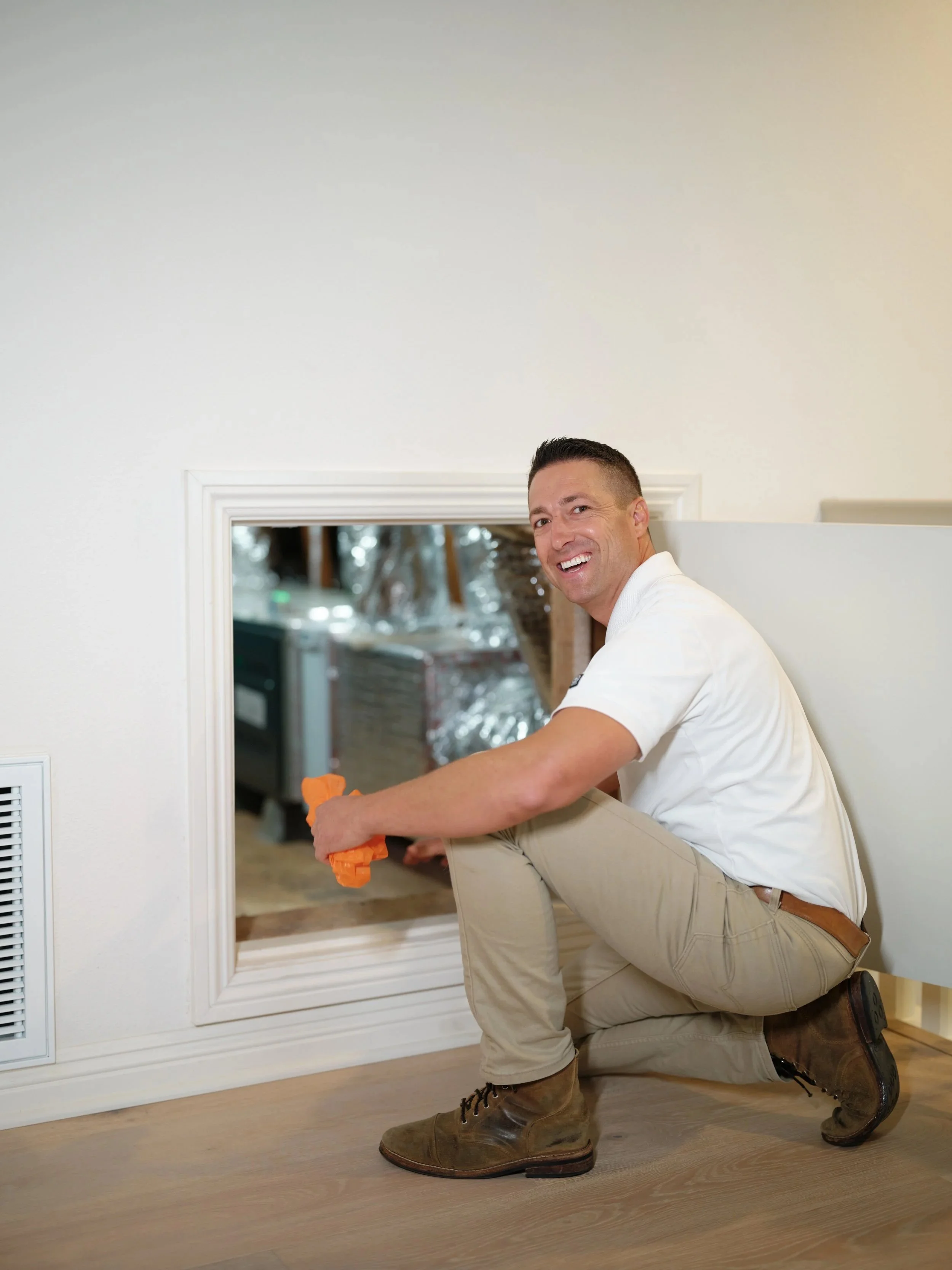 J Martin HVAC technician smiling while inspecting an attic furnace unit through an access panel during a fall heater tune-up in Orange County, wearing a white J Martin polo shirt