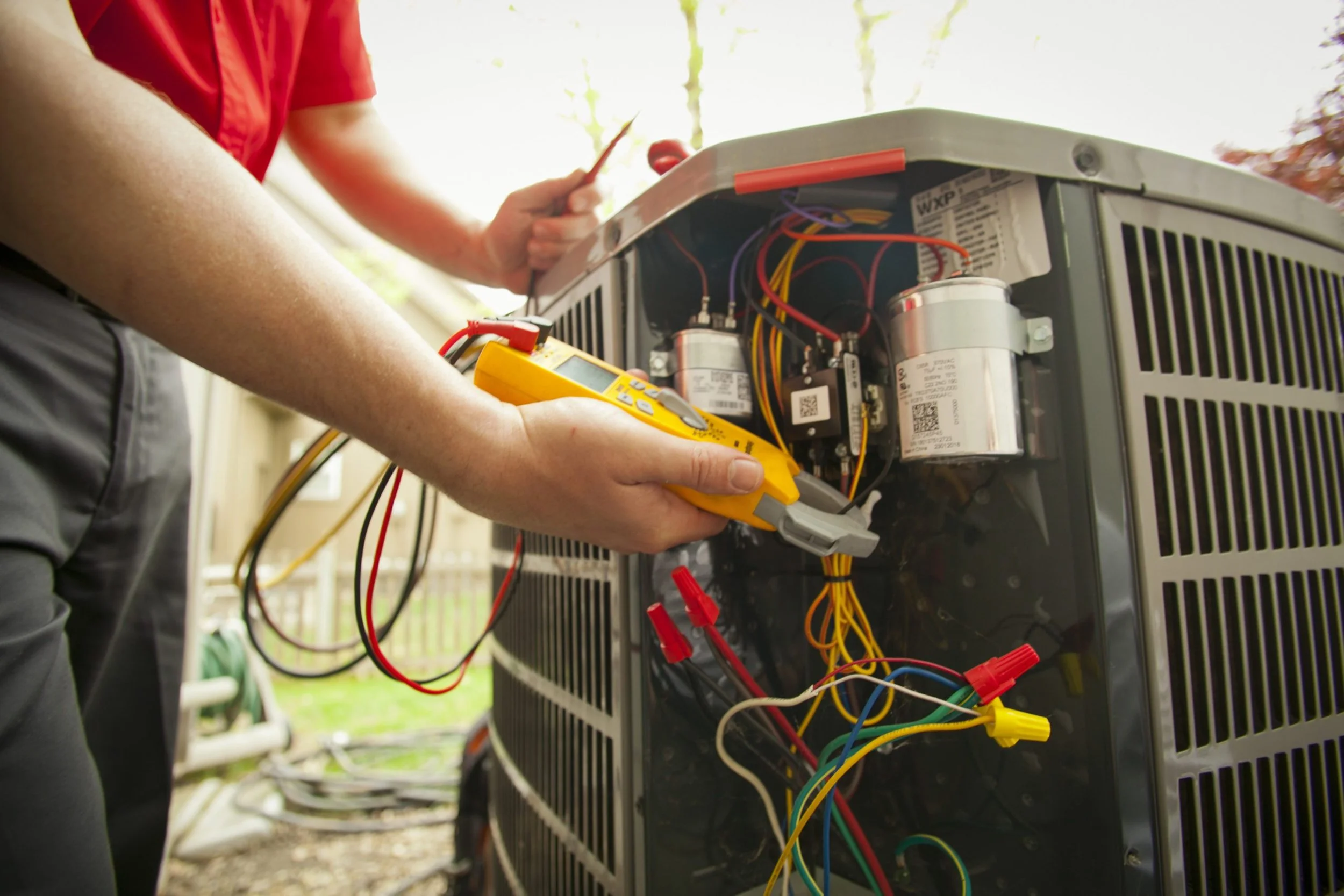 J Martin HVAC technician holding a multimeter and testing electrical components inside an open outdoor air conditioner condenser unit during a professional AC tune-up in Orange County
