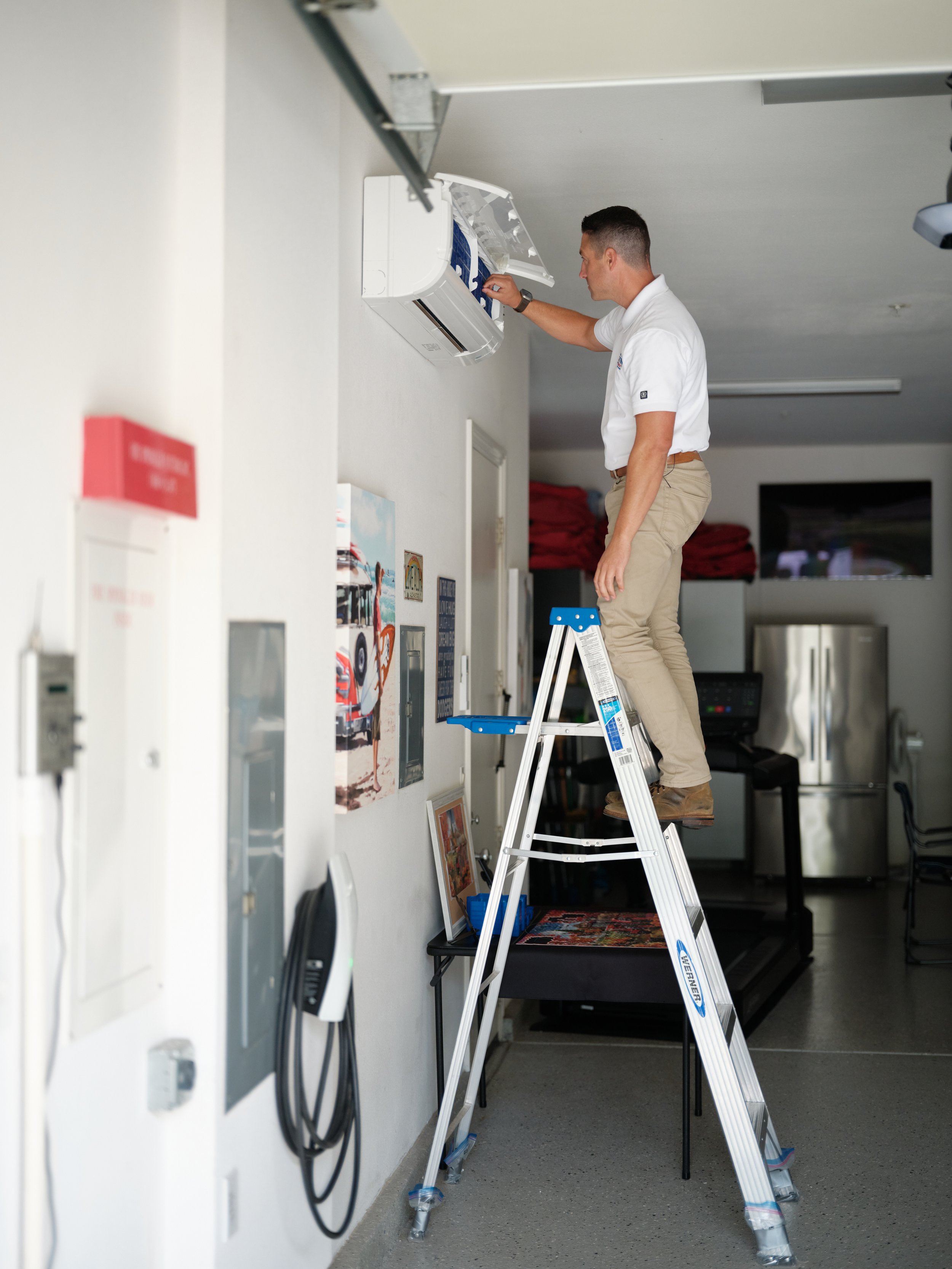 HVAC technician servicing a wall-mounted mini-split heat pump in a garage with an EV charger visible on the wall