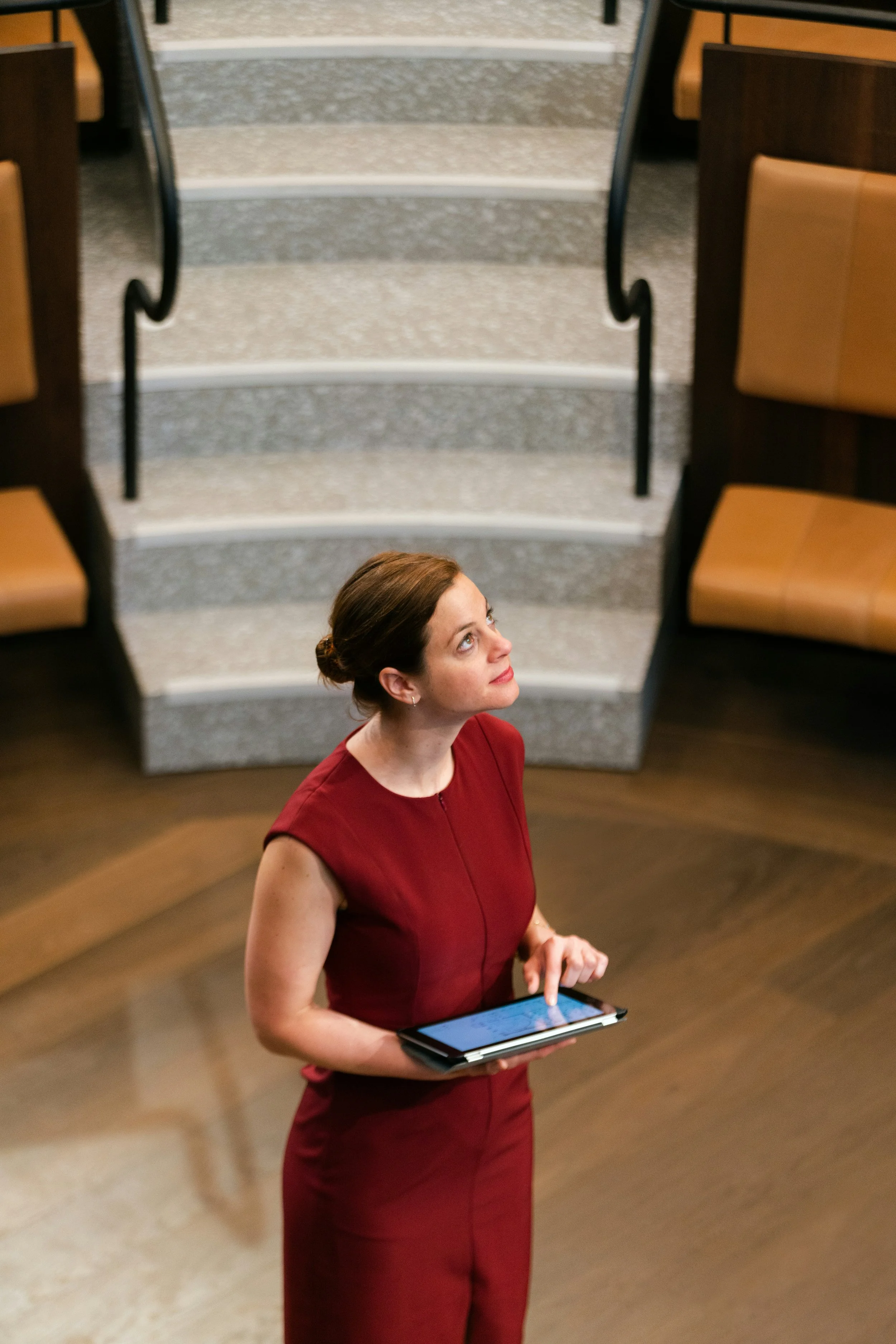 A woman with brown hair in an updo, wearing a sleeveless red dress, holding a tablet, standing in a building with wooden floors and seats, looking upward.
