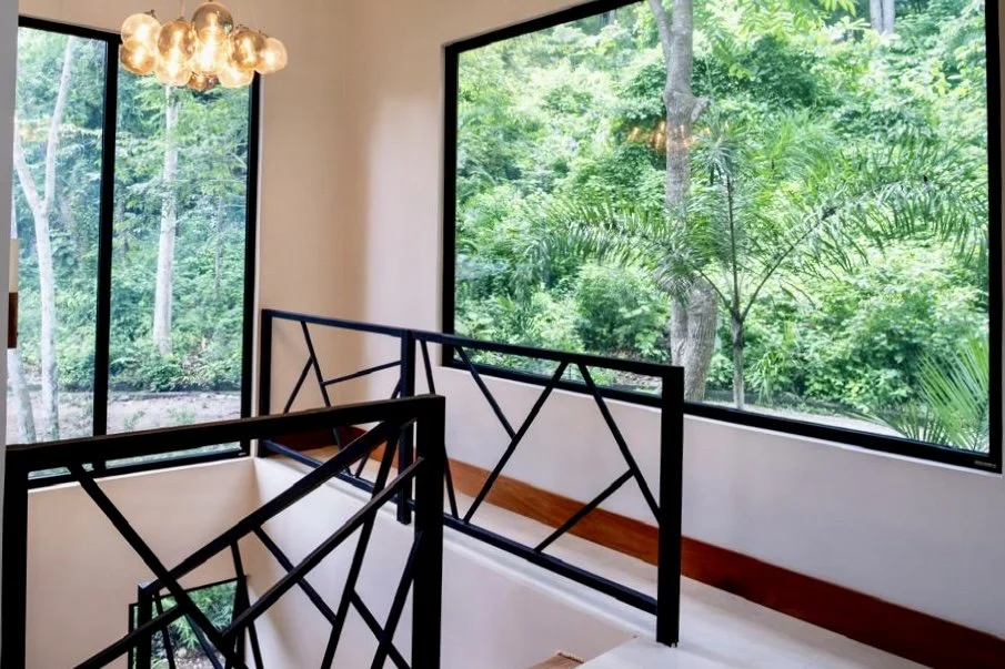 Bright upper-level landing with black metal railings, large windows and lush tropical forest views at House of Light in Paquera, Costa Rica