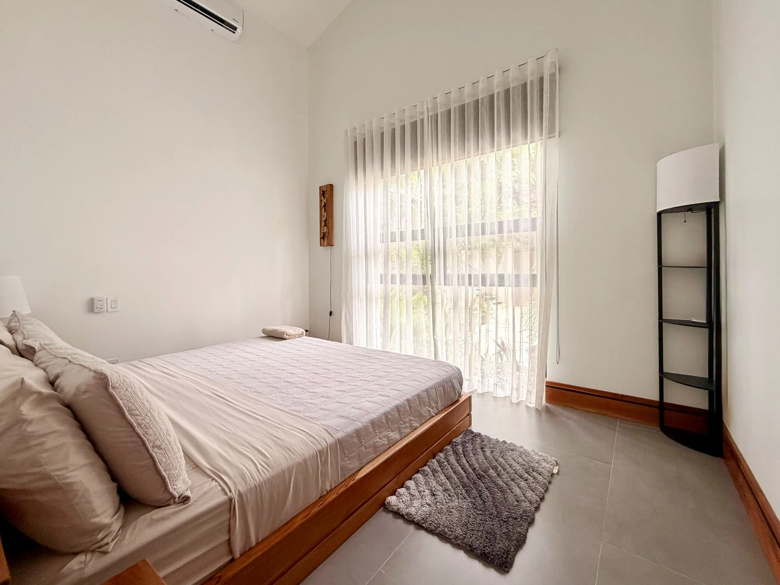 Casa Colina bright bedroom with wooden bed frame, soft neutral bedding and large window overlooking tropical greenery.
