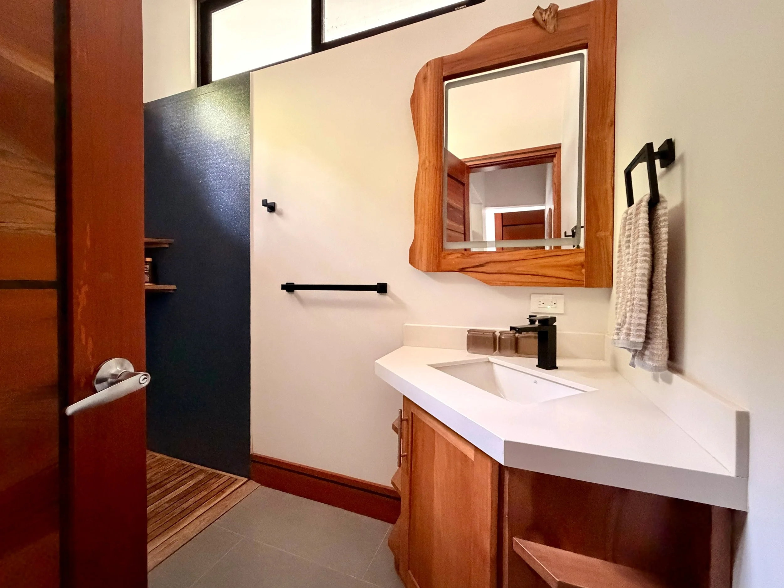 Casa Colina modern bathroom vanity with white countertop, natural wood cabinetry and decorative wooden-framed mirror.
