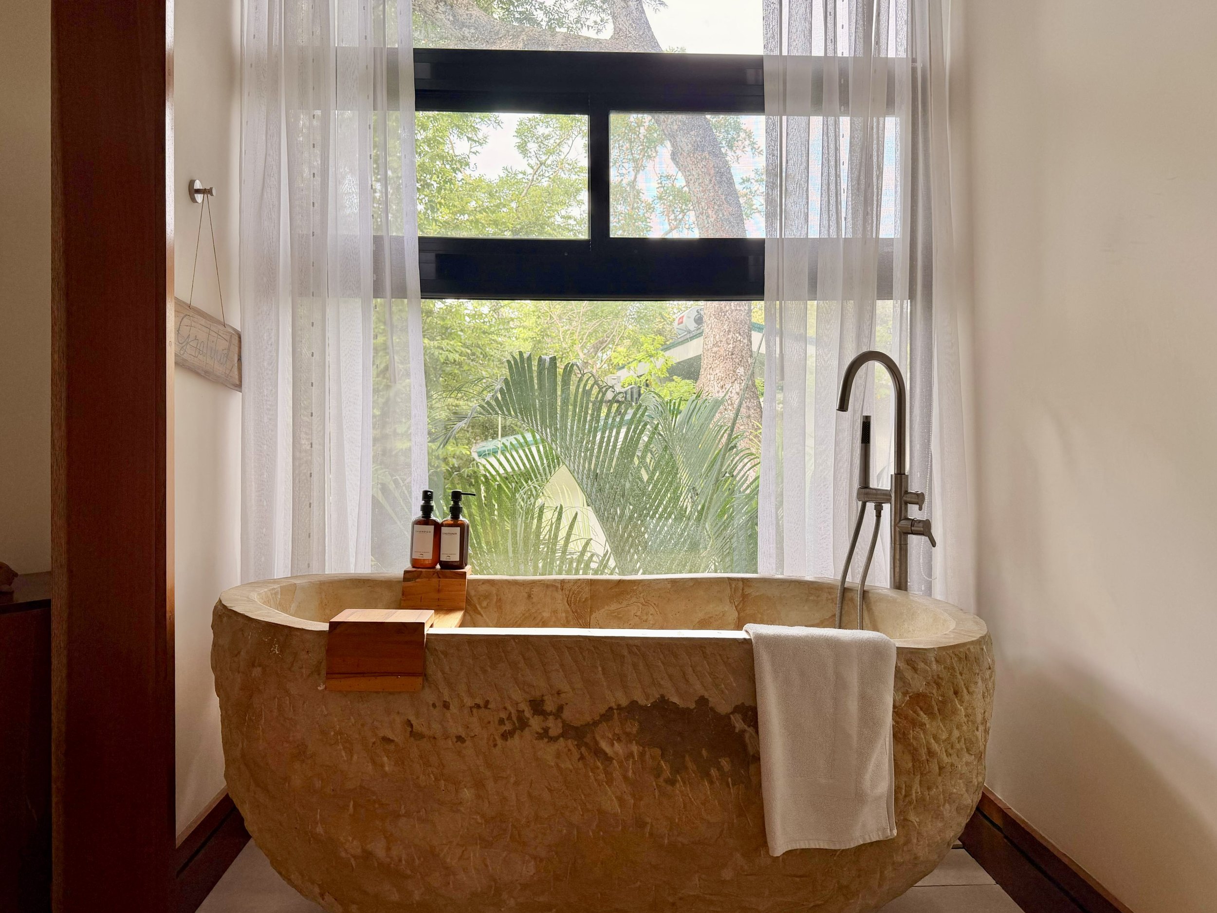 Casa Quantum hand-carved natural stone bathtub beside a large window overlooking tropical greenery.