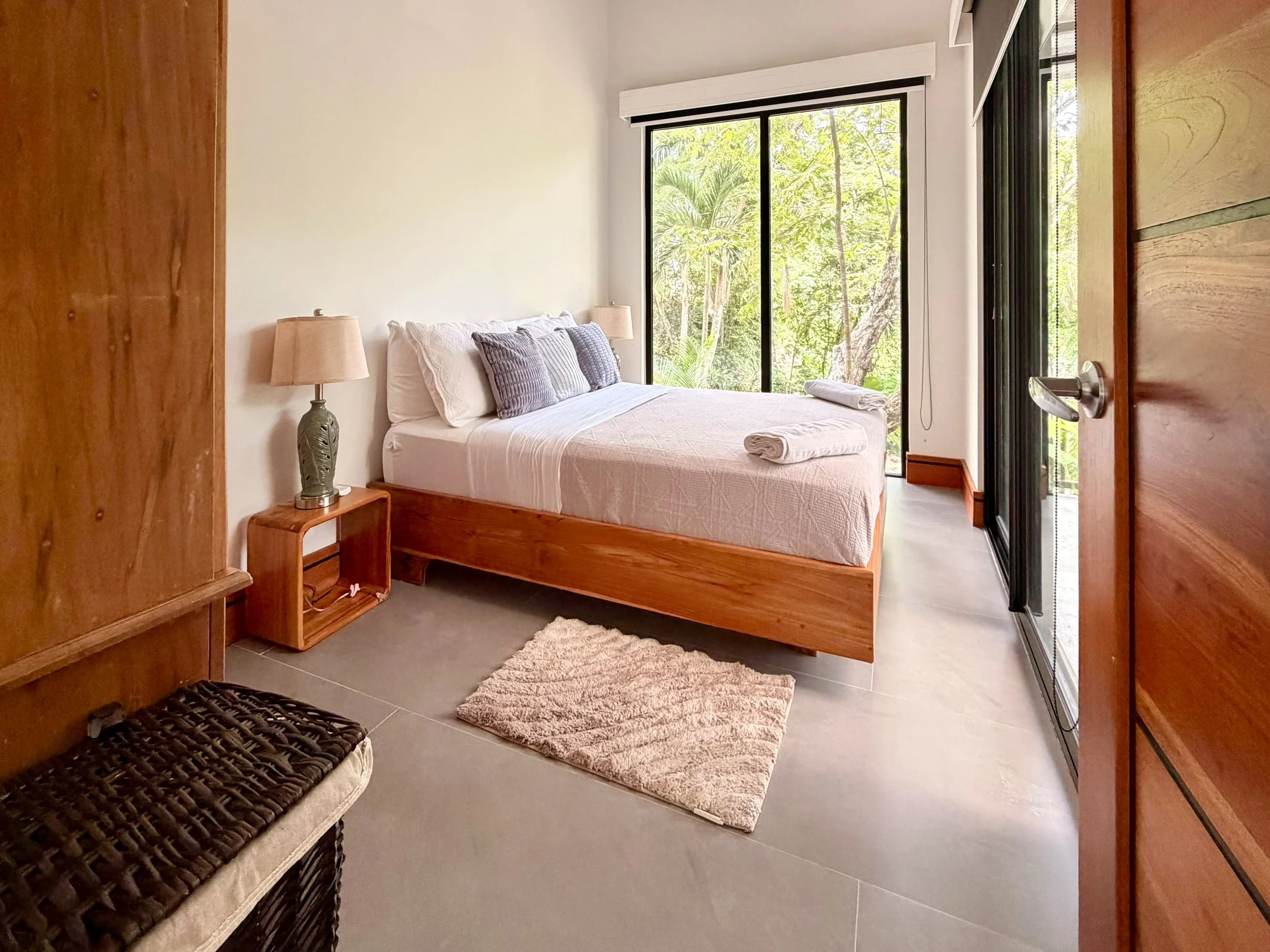 Casa Colina bright upstairs bedroom with wooden bed frame and large glass doors overlooking tropical greenery.