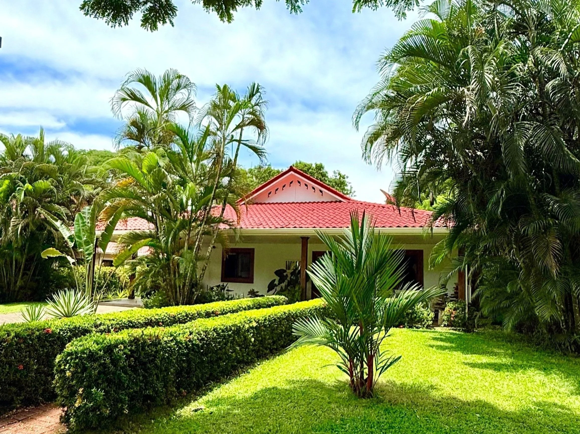 A house with a red-tiled roof surrounded by lush green tropical plants and trees, with a well-manicured lawn in the foreground and a partly cloudy sky above.