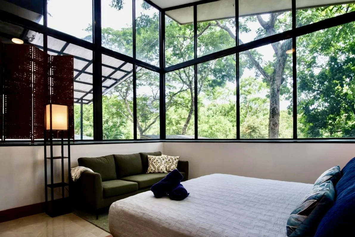 Light-filled bedroom with expansive corner windows, comfortable seating and panoramic tropical forest views at House of Light in Paquera, Costa Rica