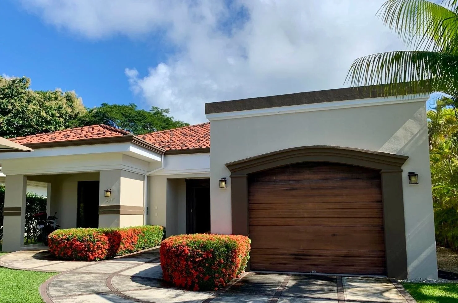 Front view of a modern house with white exterior walls, a brown wooden garage door, red tiled roof, and landscaped yard with bushes and a curved walkway, sunny sky with some clouds, and tropical plants.
