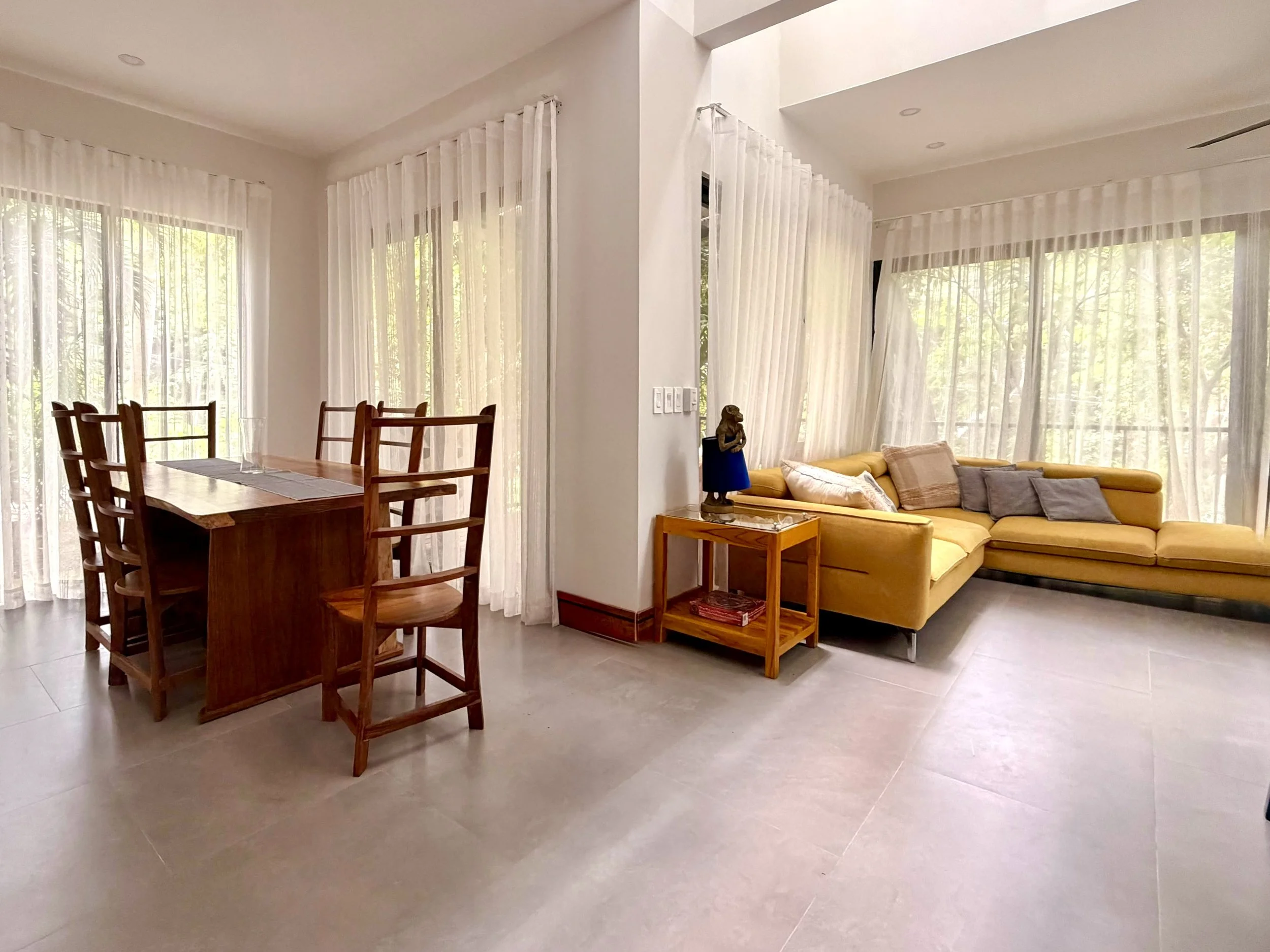 Casa Colina furnished living area with yellow sofa, dining furniture and large windows overlooking tropical greenery.