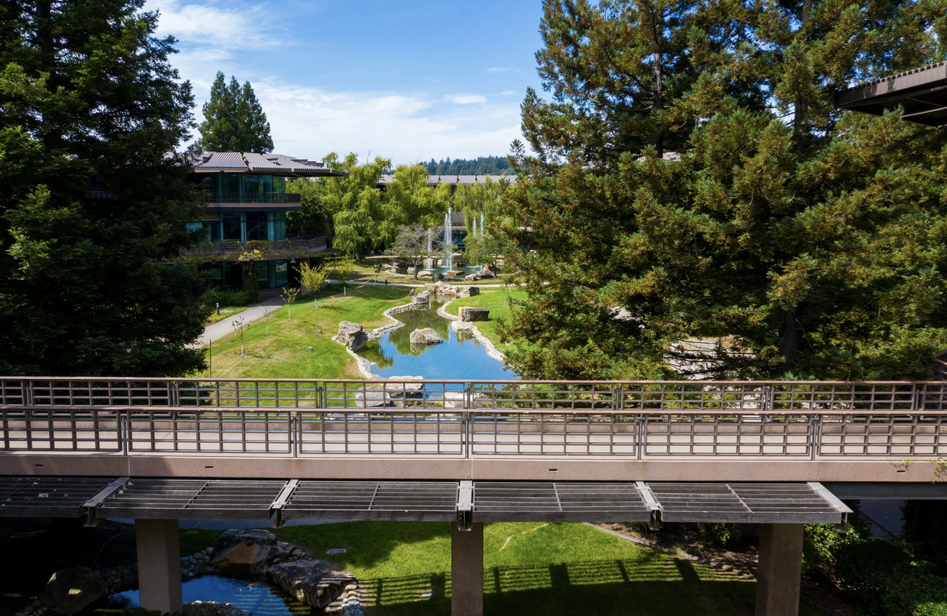 View from a balcony overlooking a landscaped area with a small stream, trees, and a fountain, with buildings in the background under a partly cloudy sky.