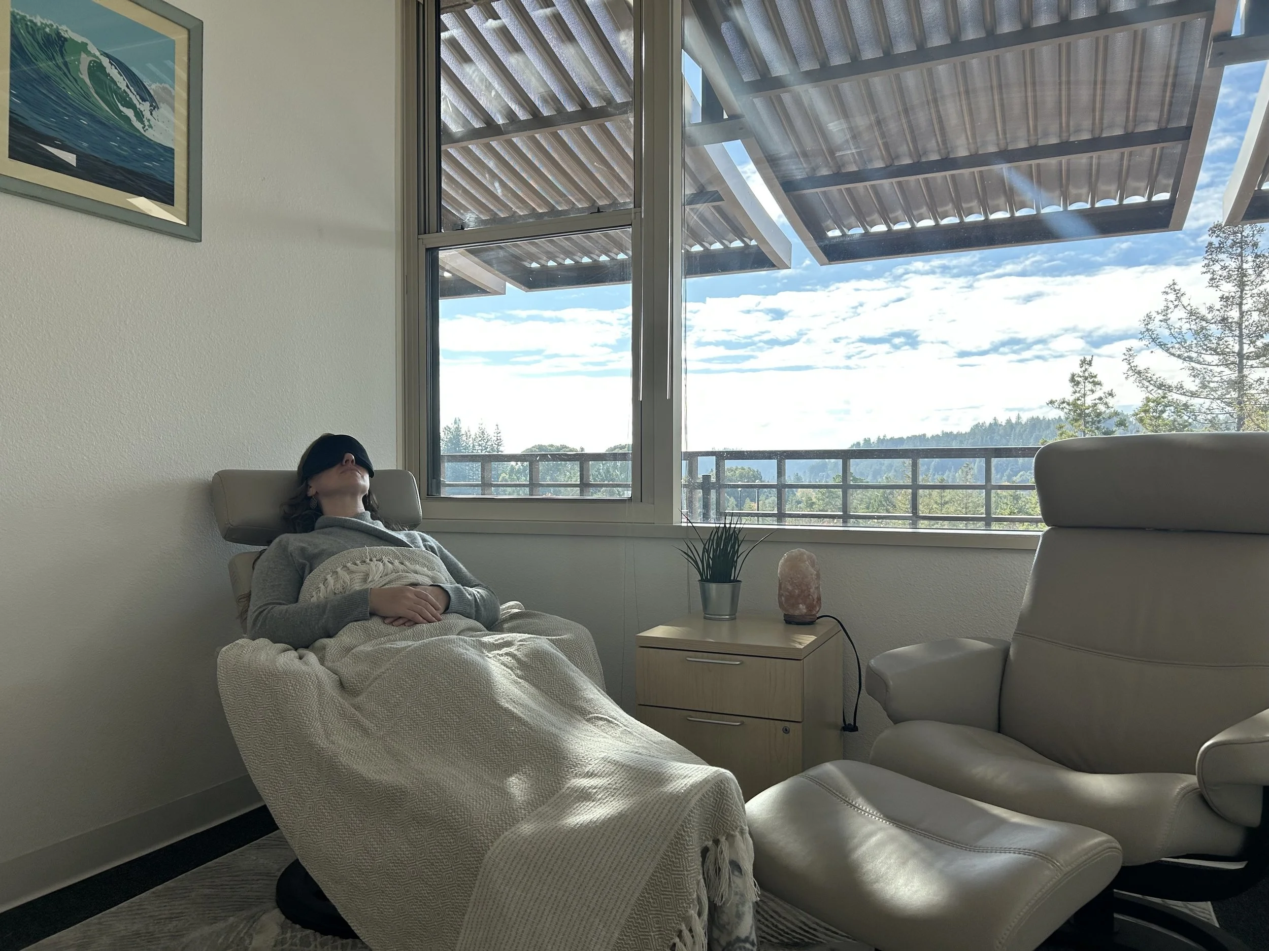 A woman wearing a sleep mask relaxes in a reclining chair next to a large window showing a landscape with trees and a cloudy sky. There is a small table with a plant, salt lamp, and a cream-colored chair and ottoman nearby.