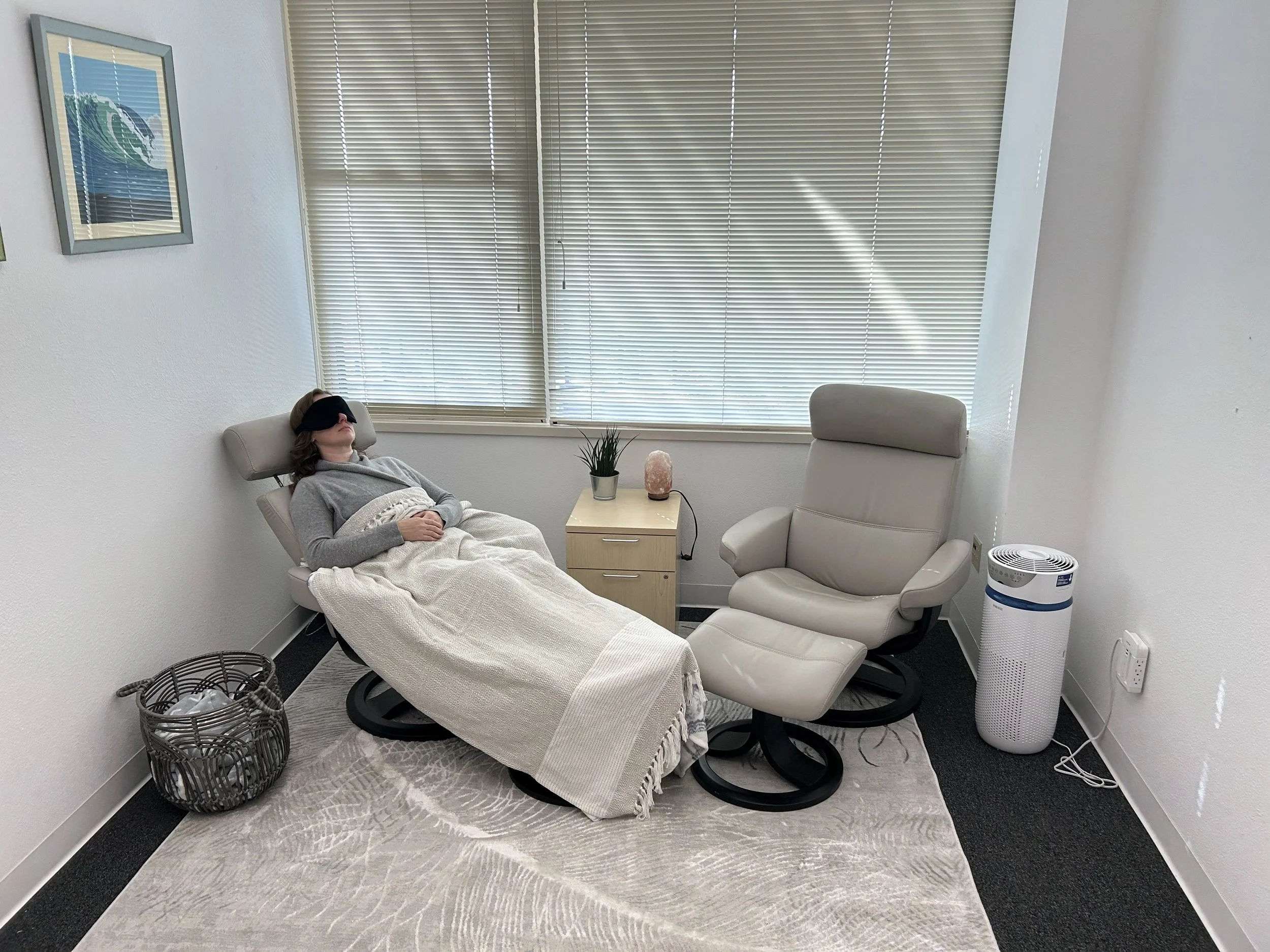 A woman lying on a reclining chair with a blanket, wearing a sleep mask, in a therapy or relaxation room with a small table, potted plant, salt lamp, air purifier, and artwork on the wall.