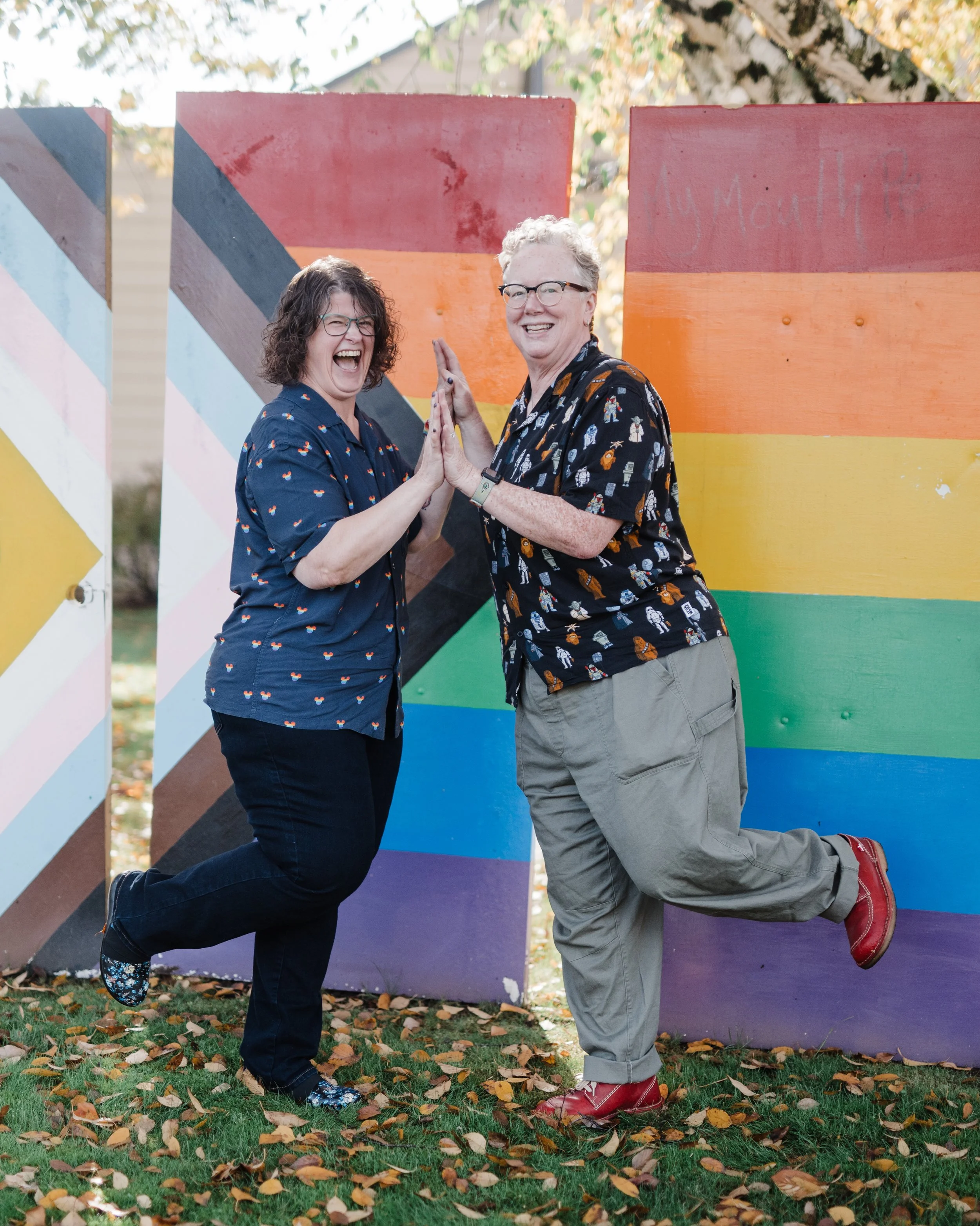 Rhonda and her wife are standing in front of a Progress Pride flag and smiling