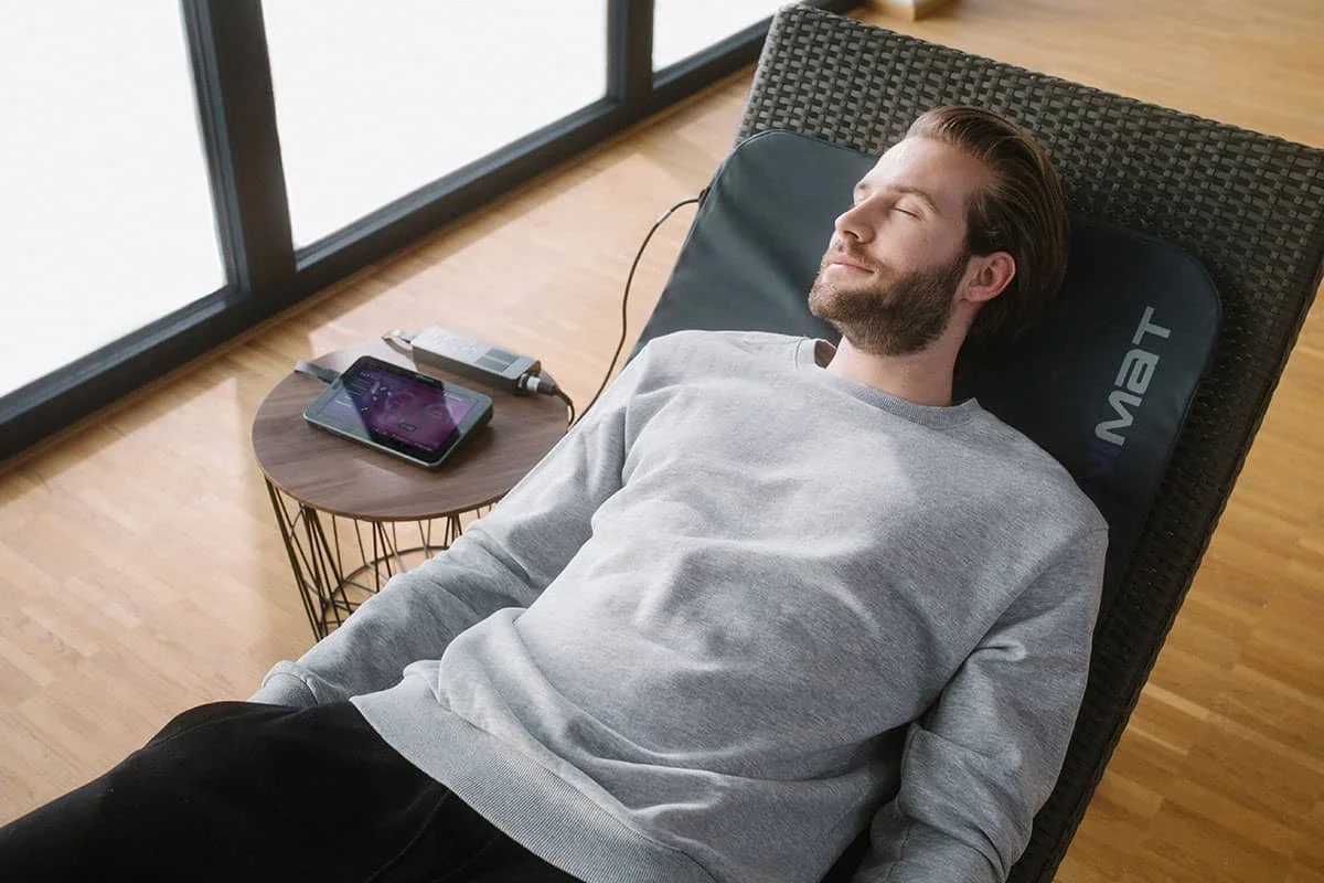 A man during PEMF therapy session with his eyes closed on a lounge chair with a neck pillow in a room with a wooden floor. There is a small round table beside him with a PEMF tablet on it. Large windows are in the background.