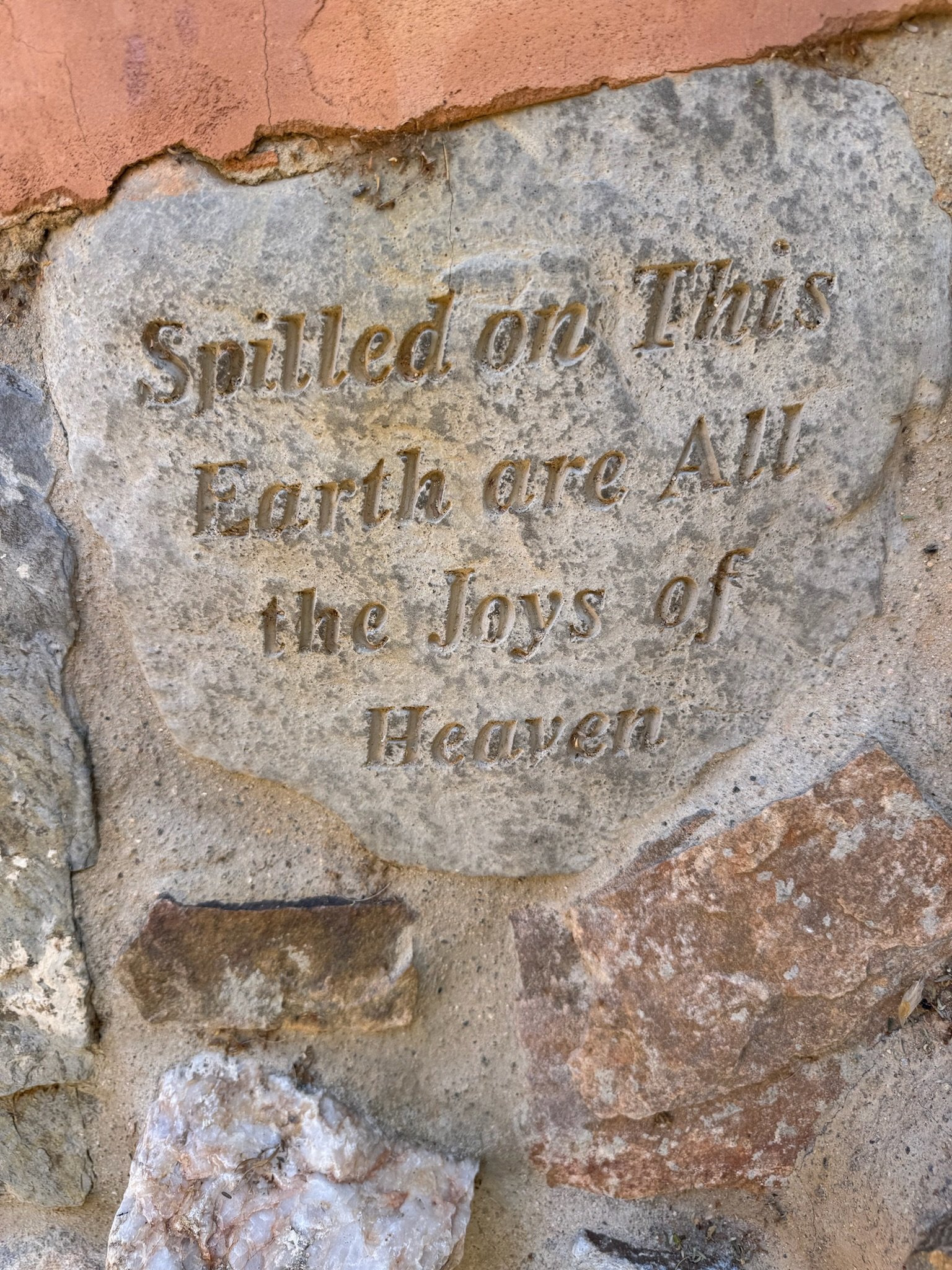 A stone plaque embedded in a wall with an engraved quote that reads, 'Spilled on This Earth are All the Joys of Heaven.'
