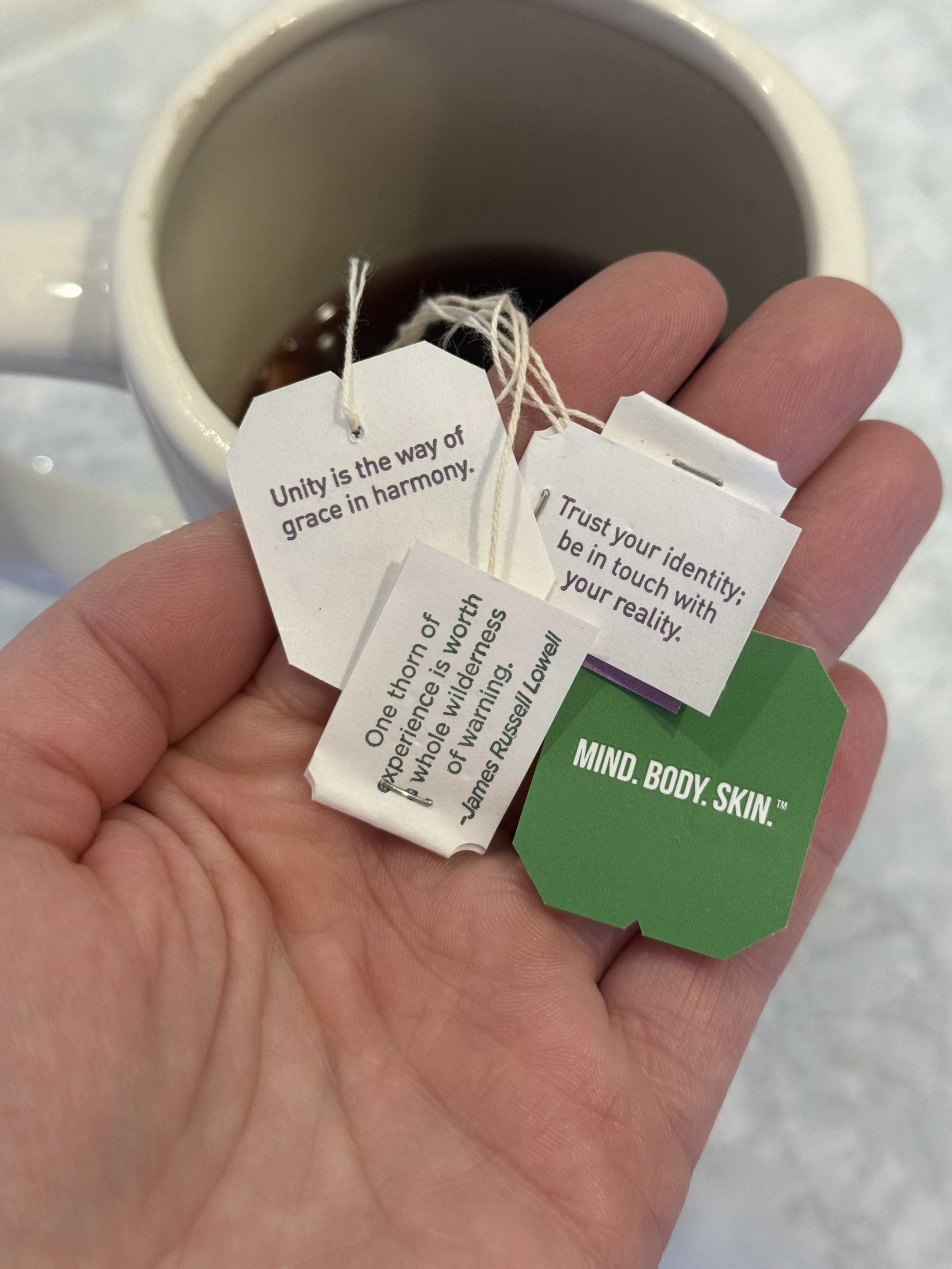 Hand holding tea bag tags and a cup of tea in the background.