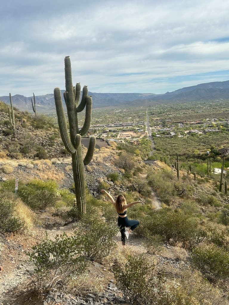 A woman practicing yoga on a trail in a desert landscape with large cactus plants, overlooking a city in the distance under a cloudy sky.