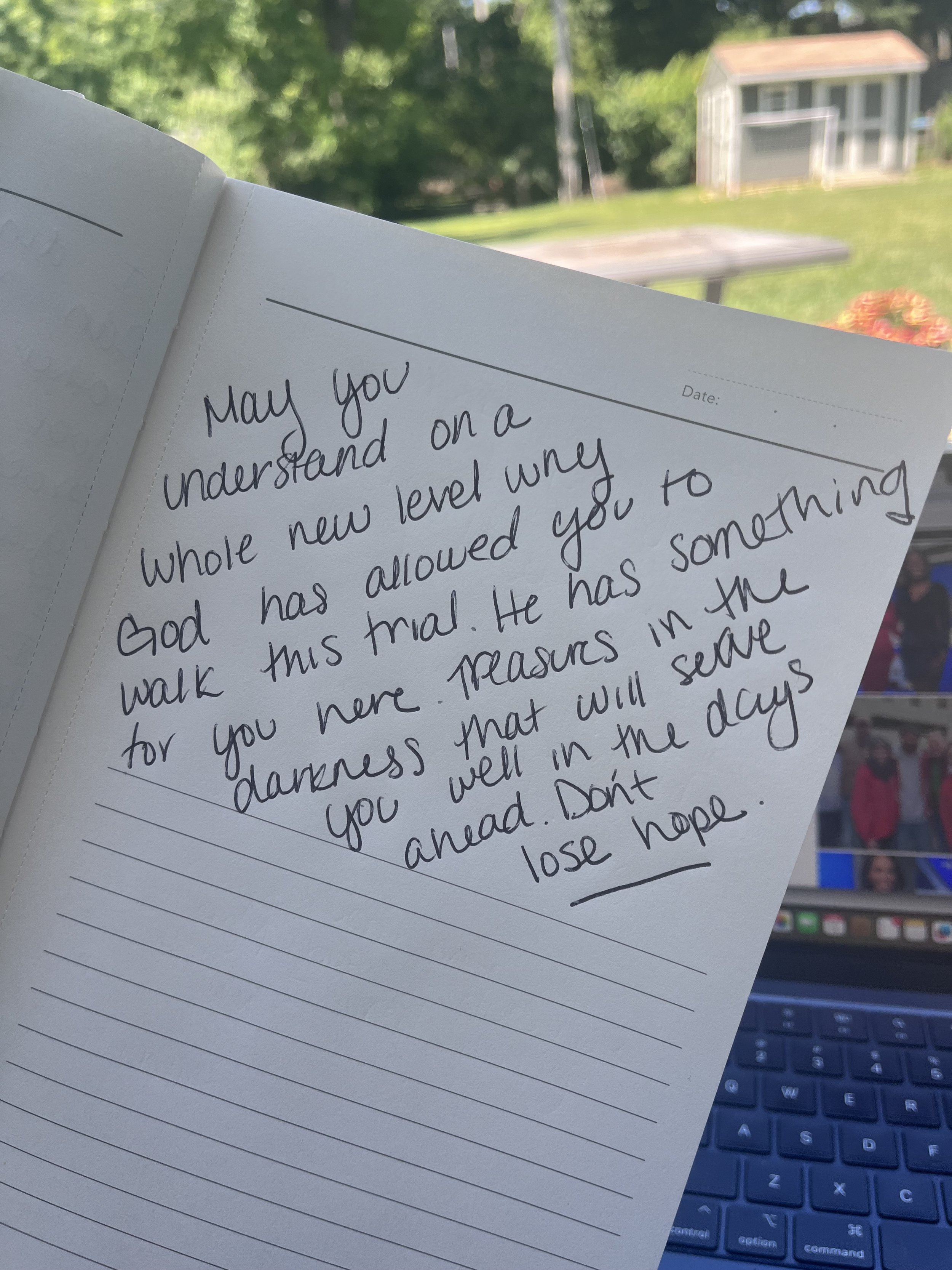 A handwritten inspirational note in a notebook with a background of a green outdoor yard, a shed, and a laptop computer.
