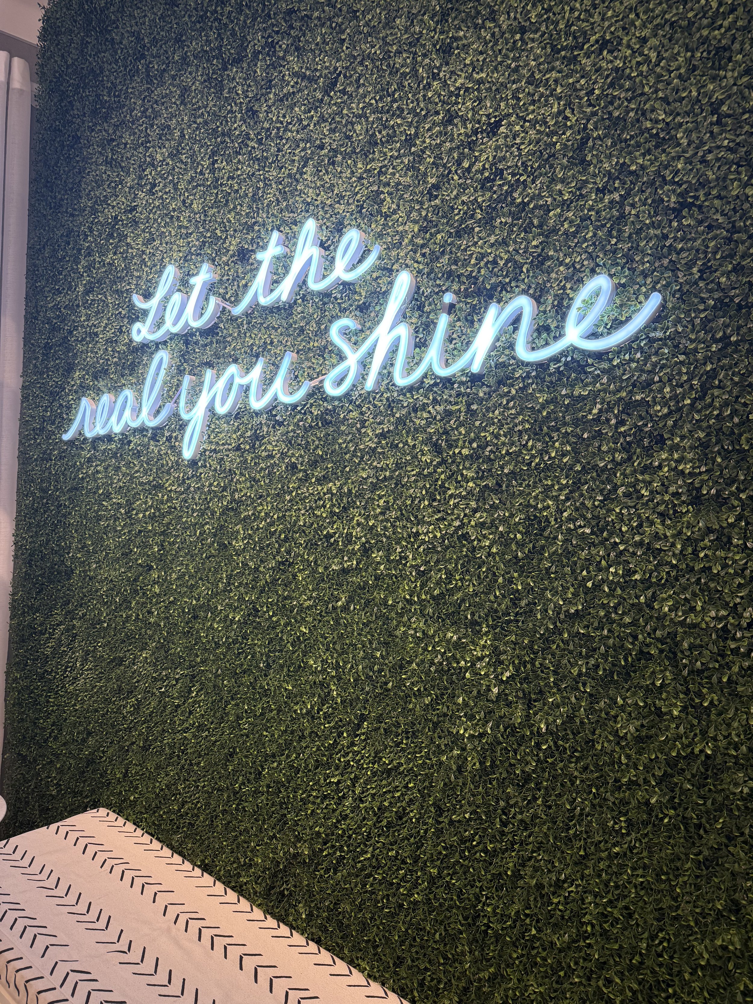 Neon sign on a green leafy wall that reads 'Let the real you shine' in cursive script.