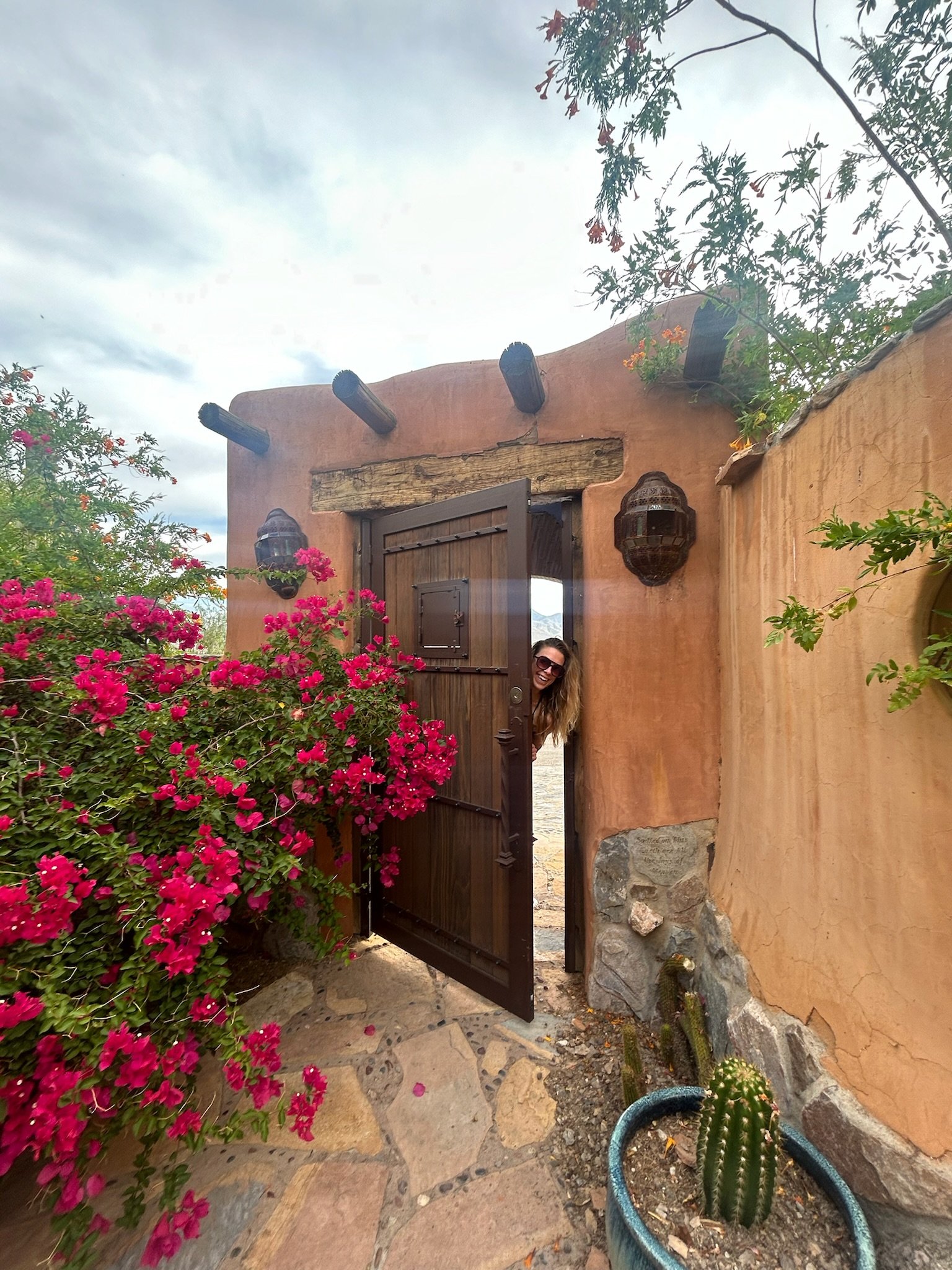 A woman peeking through an open wooden door in an adobe-style building, surrounded by pink flowers and desert plants. The sky is partly cloudy.