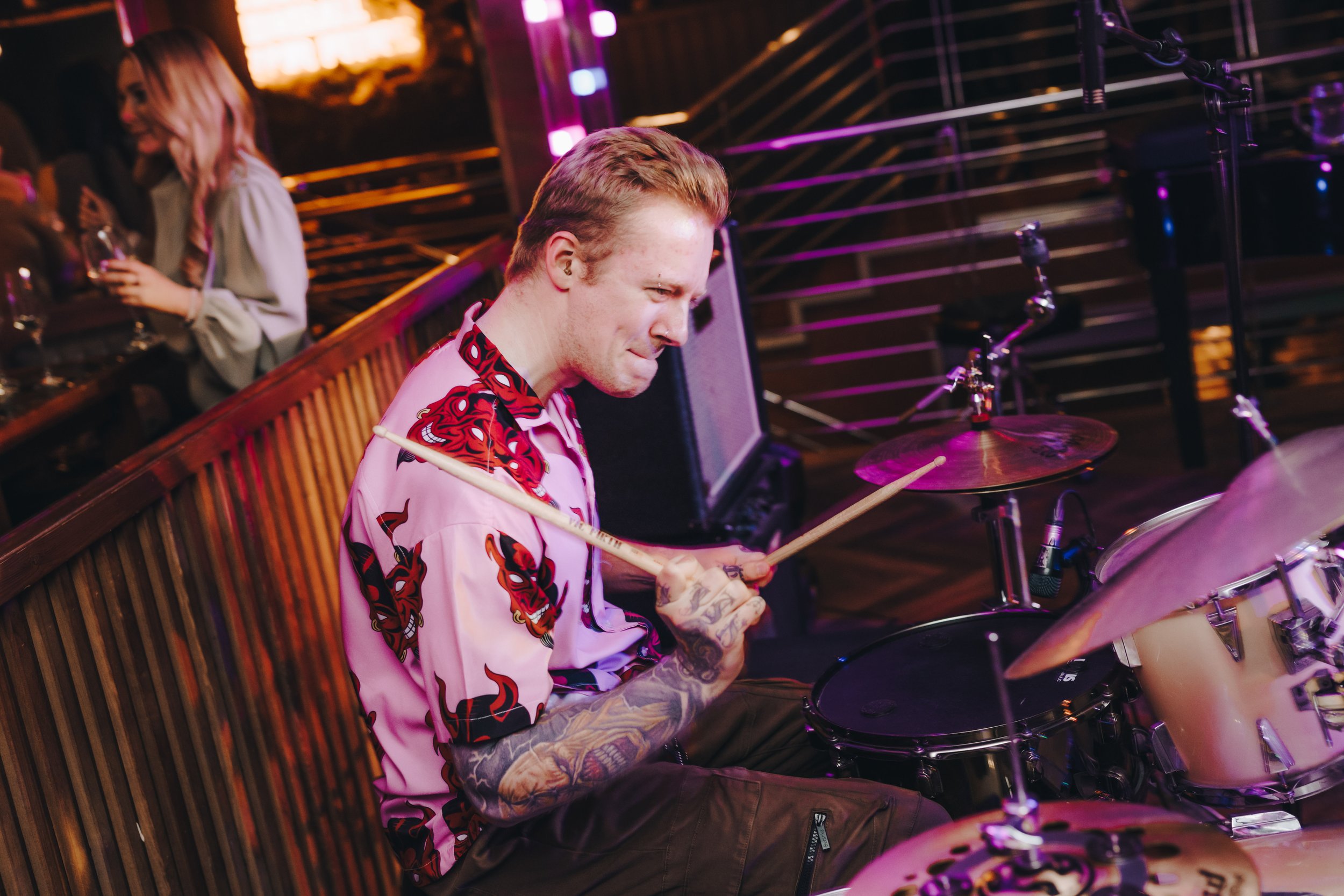 Tattooed drummer playing a drum set at a live performance with a person in the background holding a drink. Brewhemia