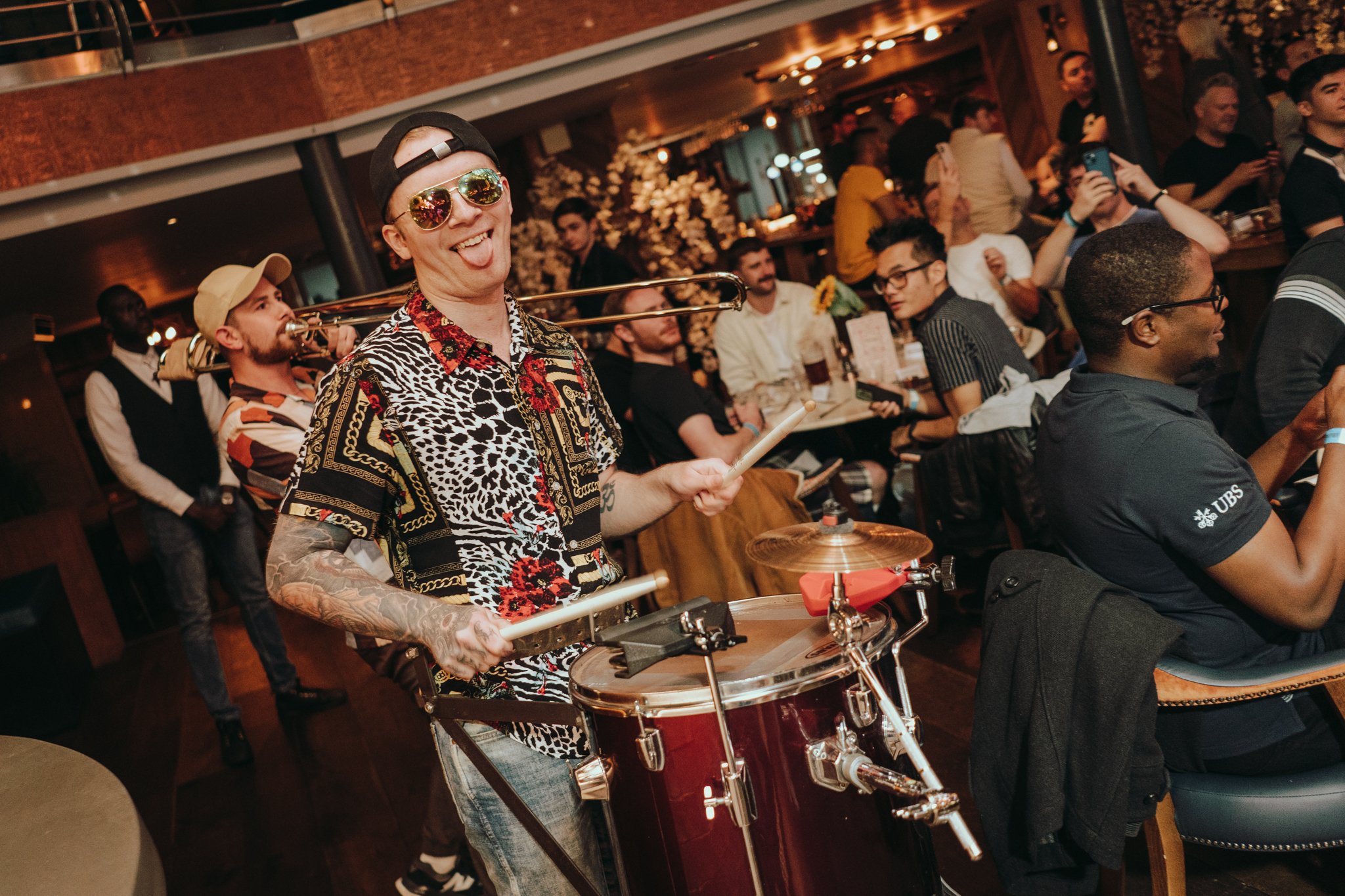 Smiling man playing percussion at a lively bar called Brewhemia in Edinburgh, surrounded by a crowd including a trombone player and people enjoying the music.