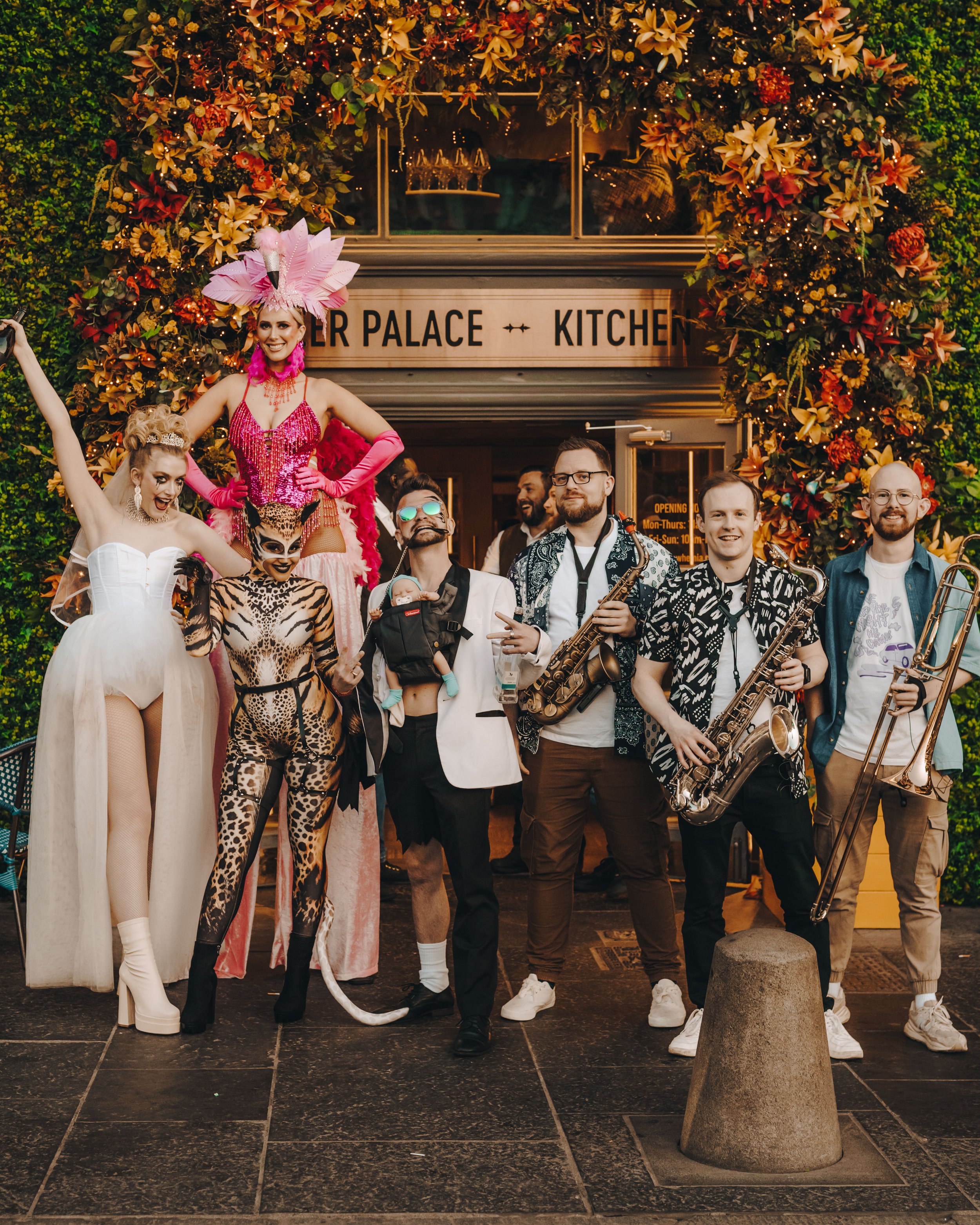 Group of performers in colorful costumes outside Brewhemia in Edinburgh, posing with musicians holding saxophones and a trombone, under a floral arch.