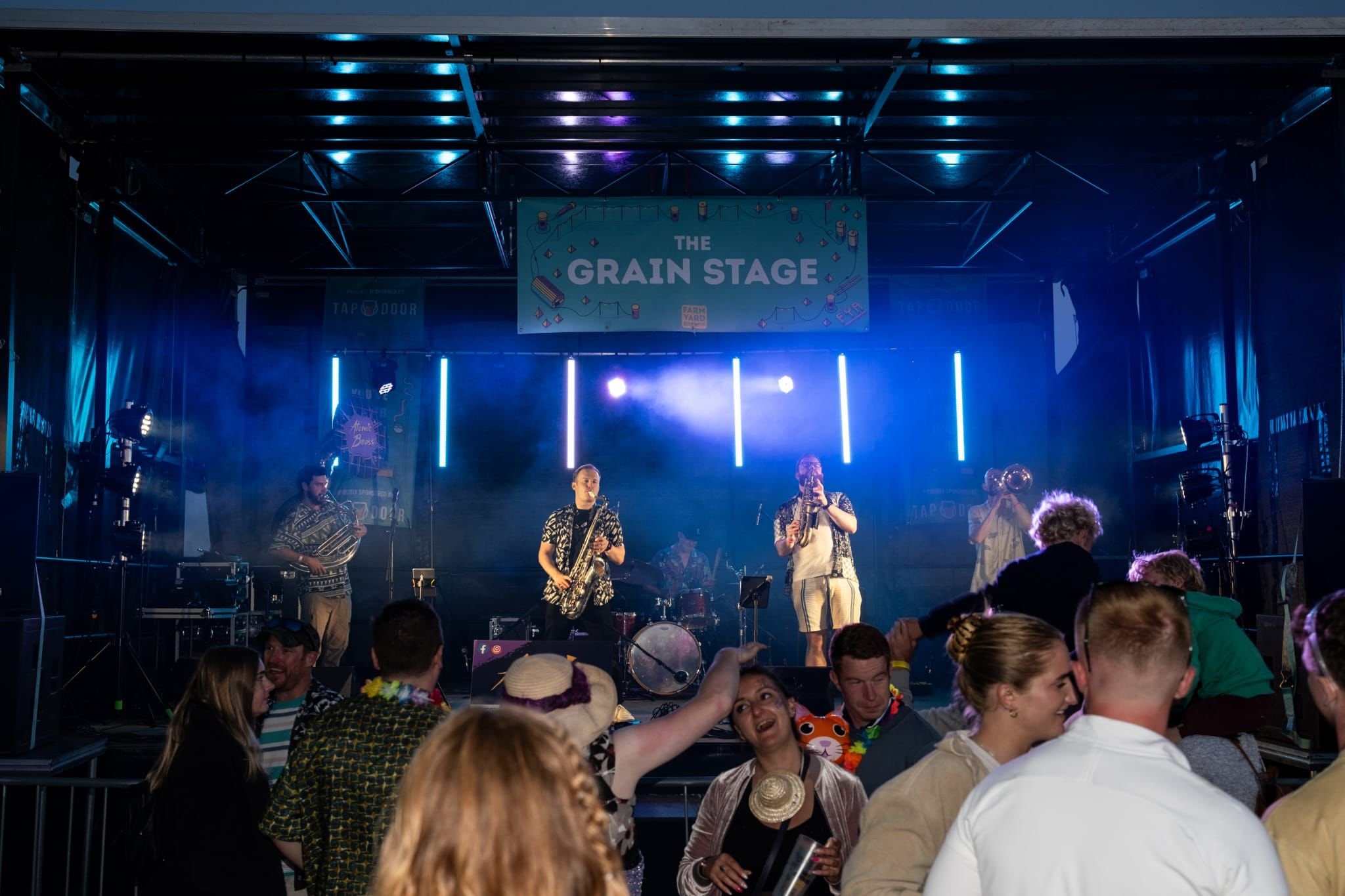 Live band performing on stage under 'The Grain Stage' sign at a music festival with people dancing in the foreground.