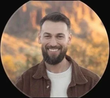 A smiling man with a beard outdoors during sunset.