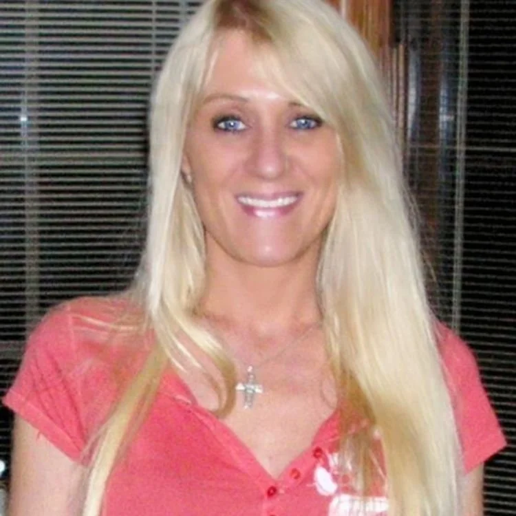 A woman with long blonde hair, wearing a pink shirt and a cross necklace, smiling at the camera.