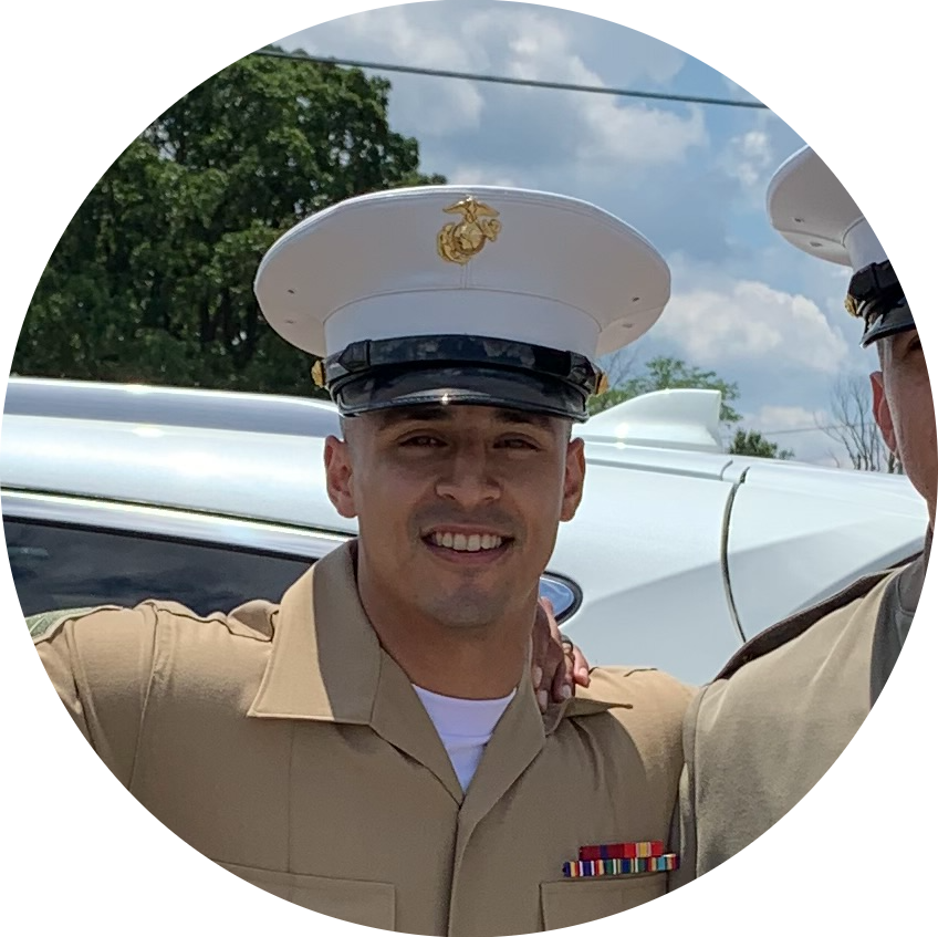 A young man in a Khaki military uniform with an American flag patch on the sleeve, smiling, wearing a white officer's cap with a gold emblem, standing outdoors next to another officer, with a white vehicle and trees in the background.