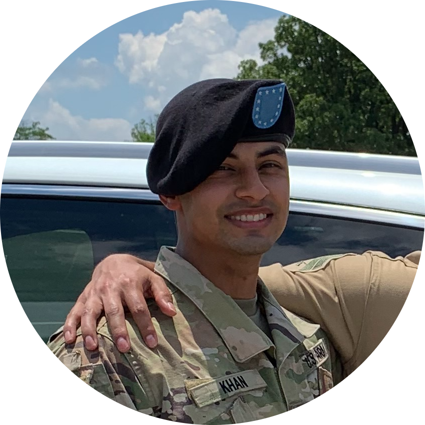 A smiling soldier in uniform with a black beret, standing outdoors next to a vehicle, with trees and a partly cloudy sky in the background.