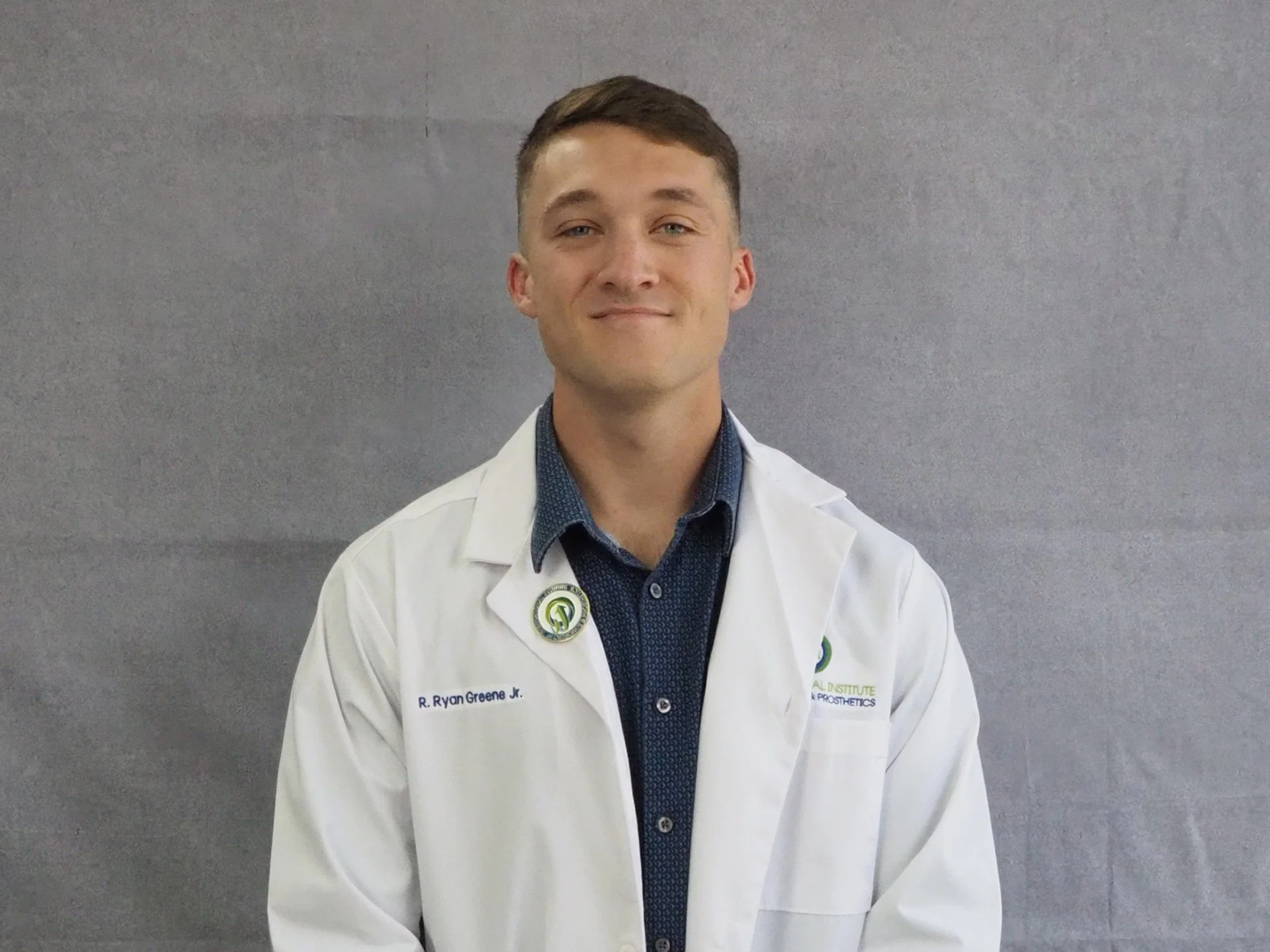 Smiling person in a white lab coat with a logo, standing against a neutral background.